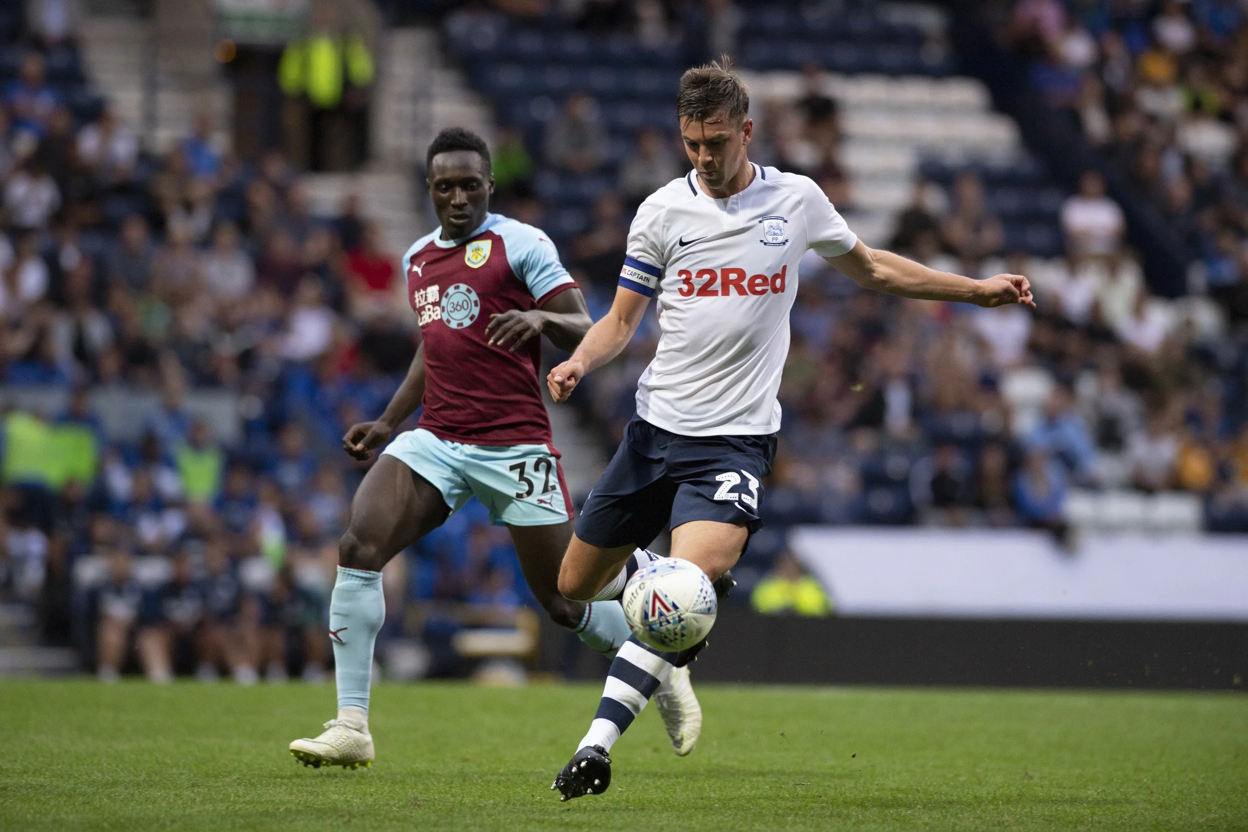 Two soccer players in action during a game, one in a white jersey and the other in a maroon and teal jersey, with a crowd in the background.