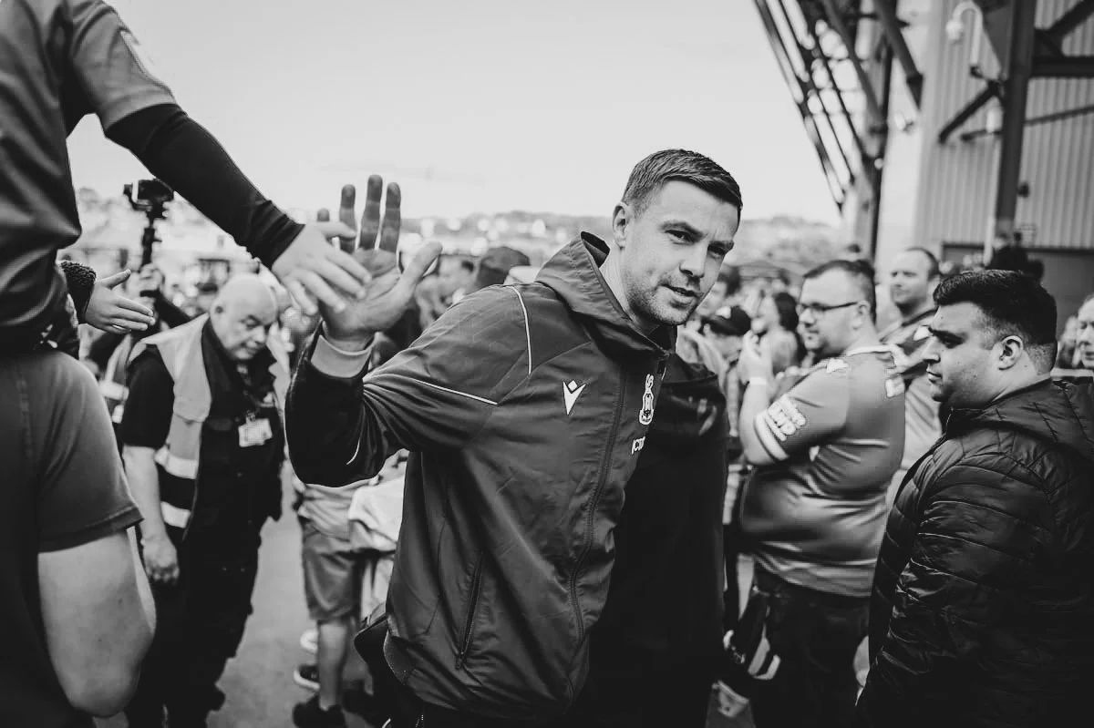 Black and white photo of a man in a sports jacket giving a handshake to someone outside the frame, surrounded by a crowd of people at or near a sports venue.