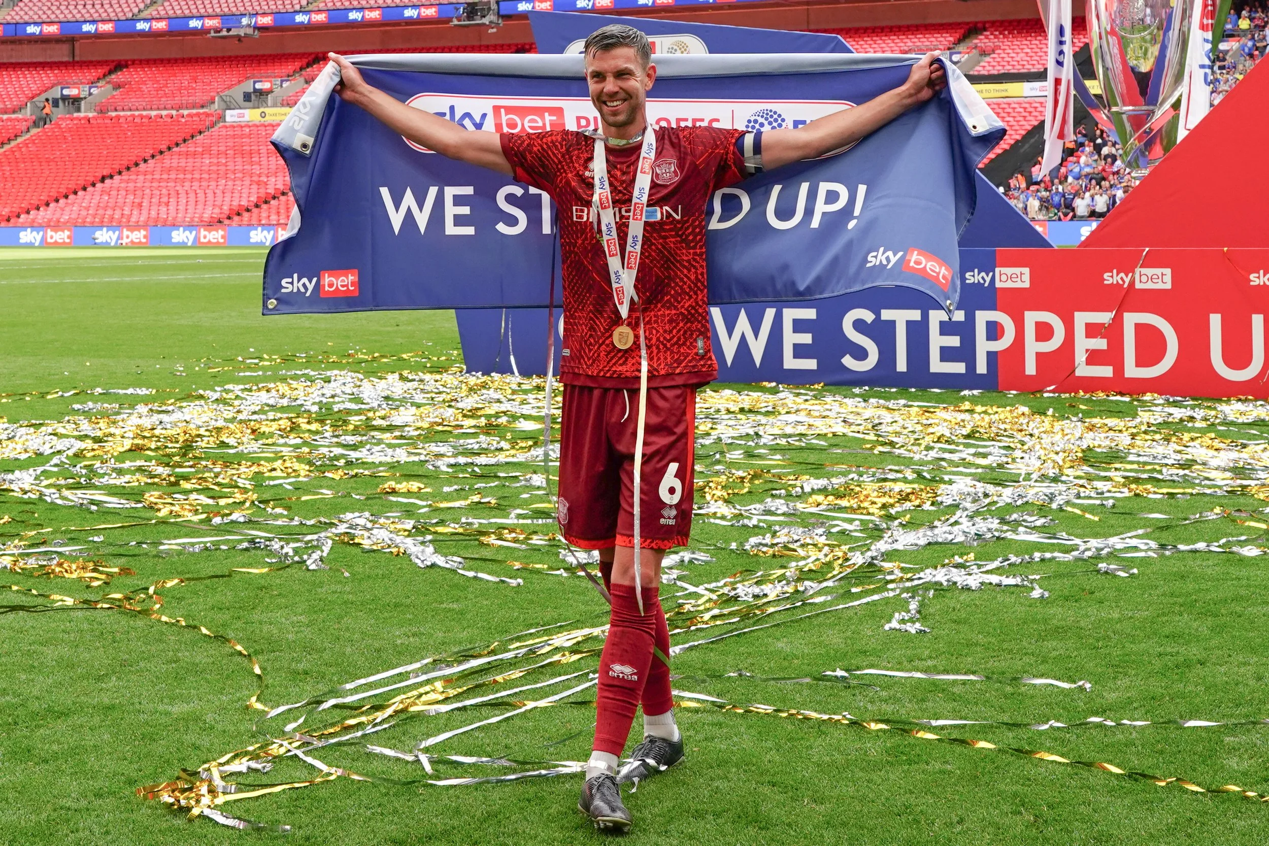 A man in a red soccer uniform holding a banner that reads 'We Step Up!' on a soccer field, with streamers and a trophy in the background, celebrating a victory.