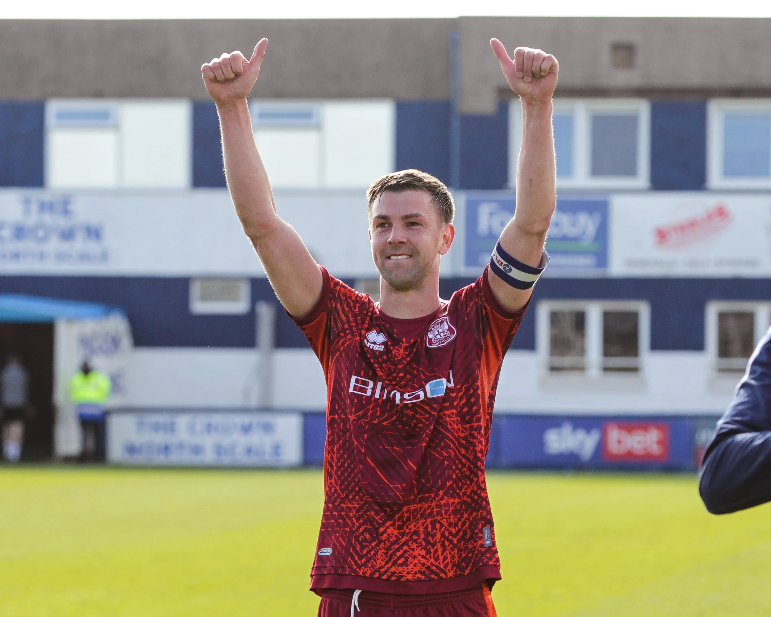 A soccer player in a maroon and red uniform with several sponsor logos, raising his arms and smiling on a soccer field.
