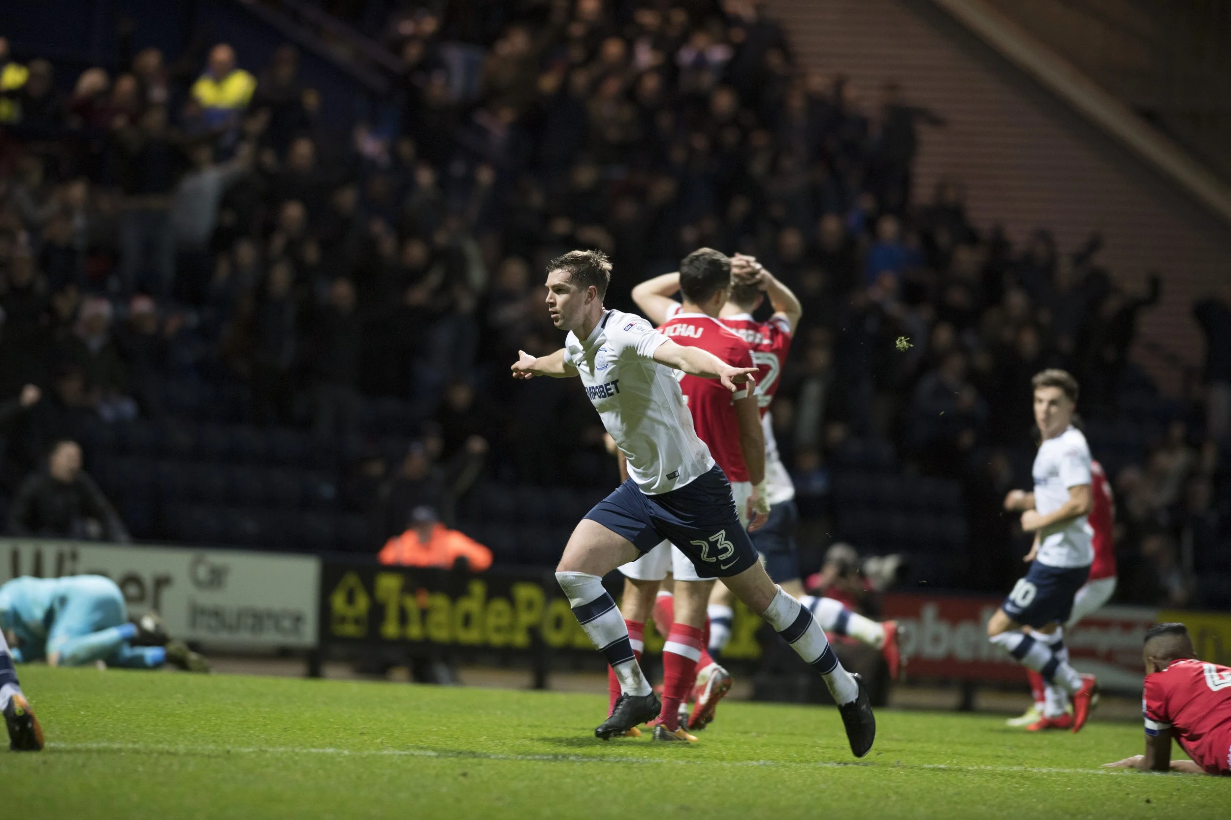 A soccer player in a white jersey with number 23 celebrates after scoring a goal during a match, with teammates and opponents visible in the background on a green field.