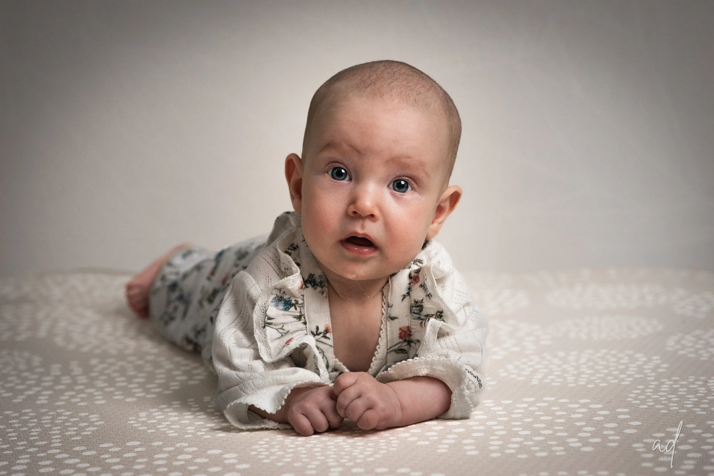 En baby med kort hår ligger på mage på en beige overflate med hvite prikker, ser rett frem med overrasket uttrykk.