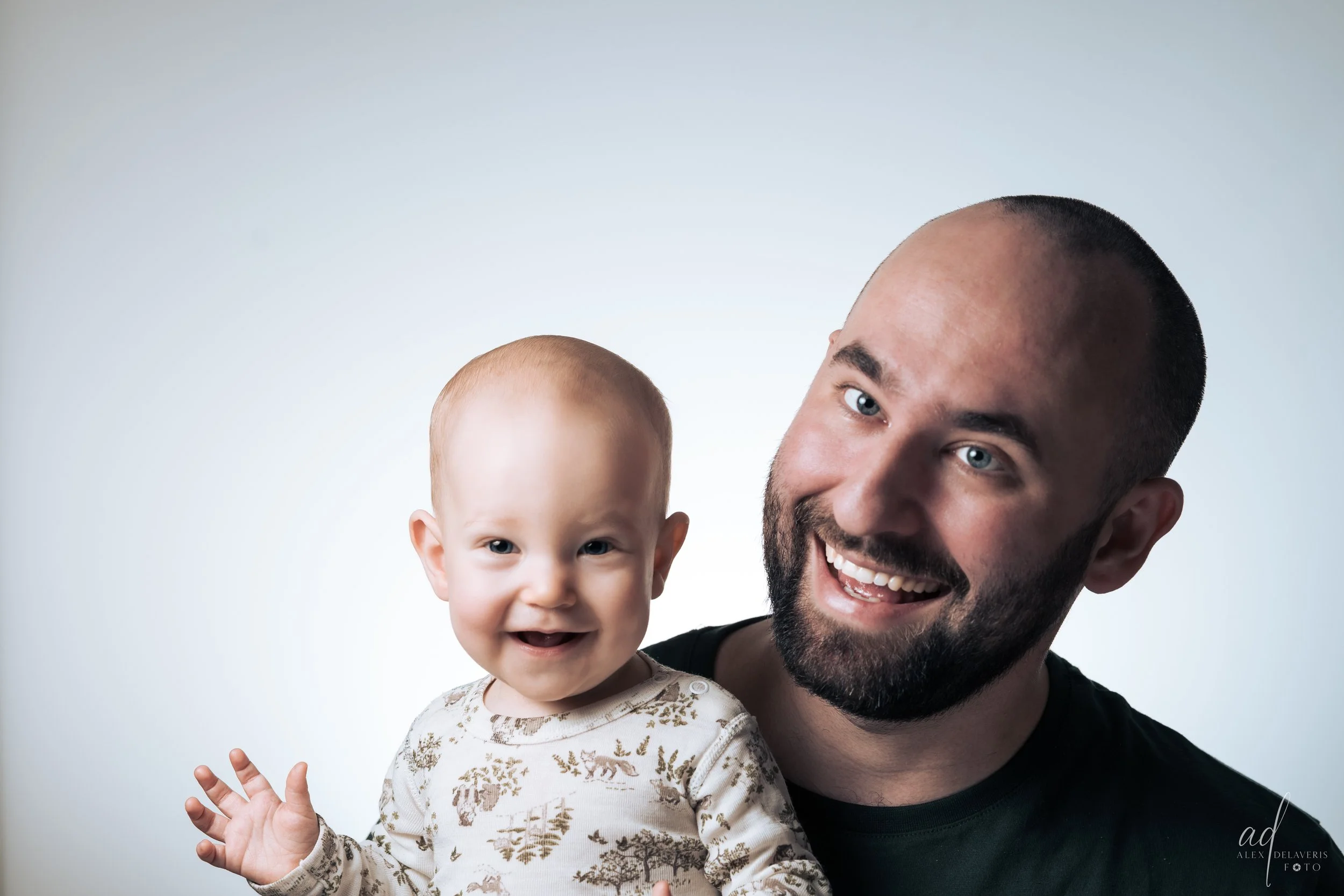 A smiling adult man with a beard holding a happy baby girl with light hair, against a plain white background.
