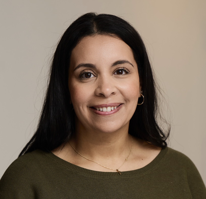 Close-up portrait of a woman with long dark hair, wearing a green top, gold hoop earrings, and a delicate gold necklace, smiling softly against a plain neutral background.