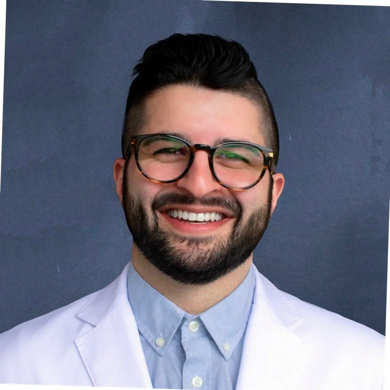 Portrait of a smiling man with dark hair, beard, and glasses, wearing a white lab coat and a light blue shirt, against a dark blue background.
