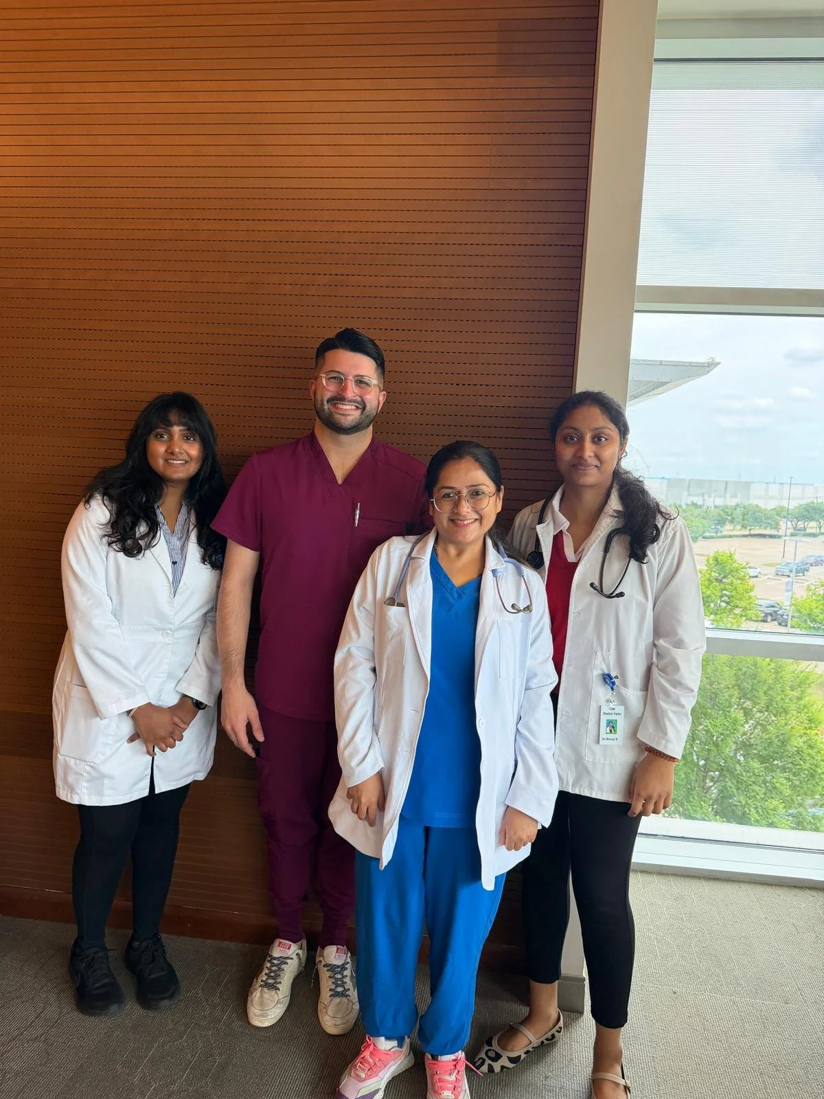 Group of five healthcare professionals, three students with Dr. Karmali, standing indoors near a window with a view of trees and the parking lot outside. They are dressed in medical scrubs and white coats, smiling.