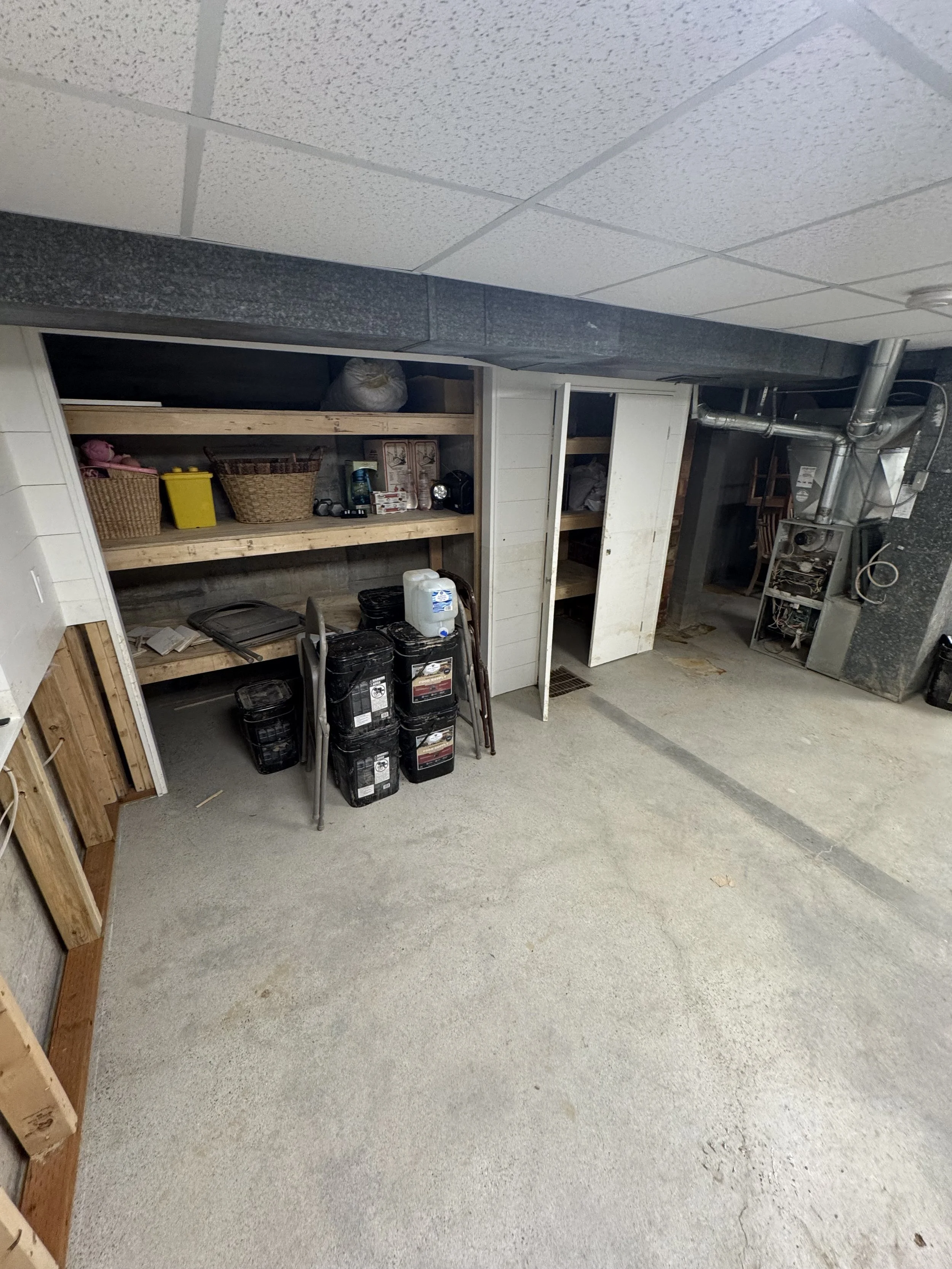 A cluttered basement storage area with wooden shelves holding bins, tools, and supplies, an open cabinet, a drain on the concrete floor, and exposed ductwork.