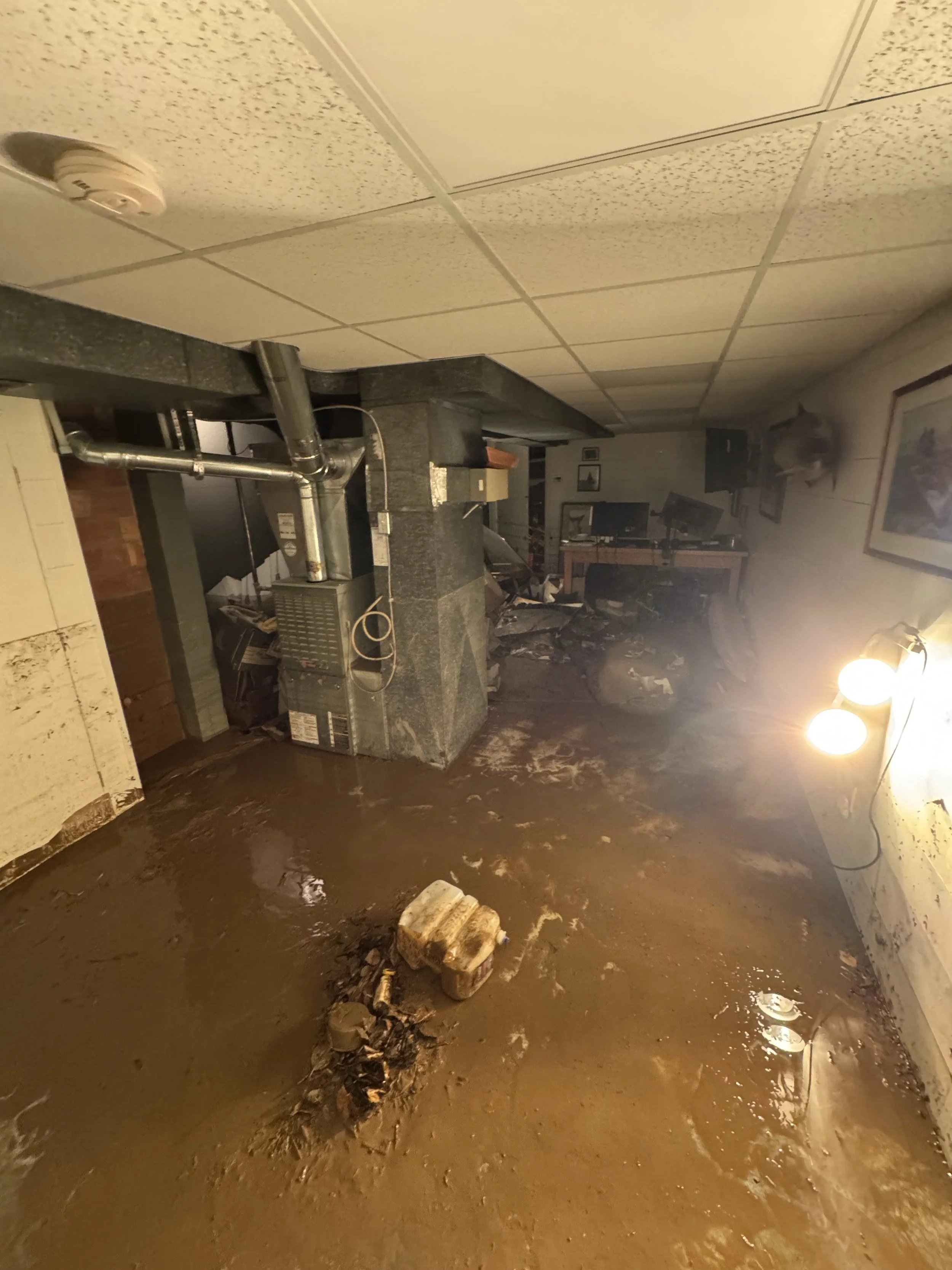 Flooded basement with muddy water covering the floor, debris, and a damaged wall. A pipe and furnace are visible on the left, and a desk with computers is in the background. Bright lights illuminate the room.
