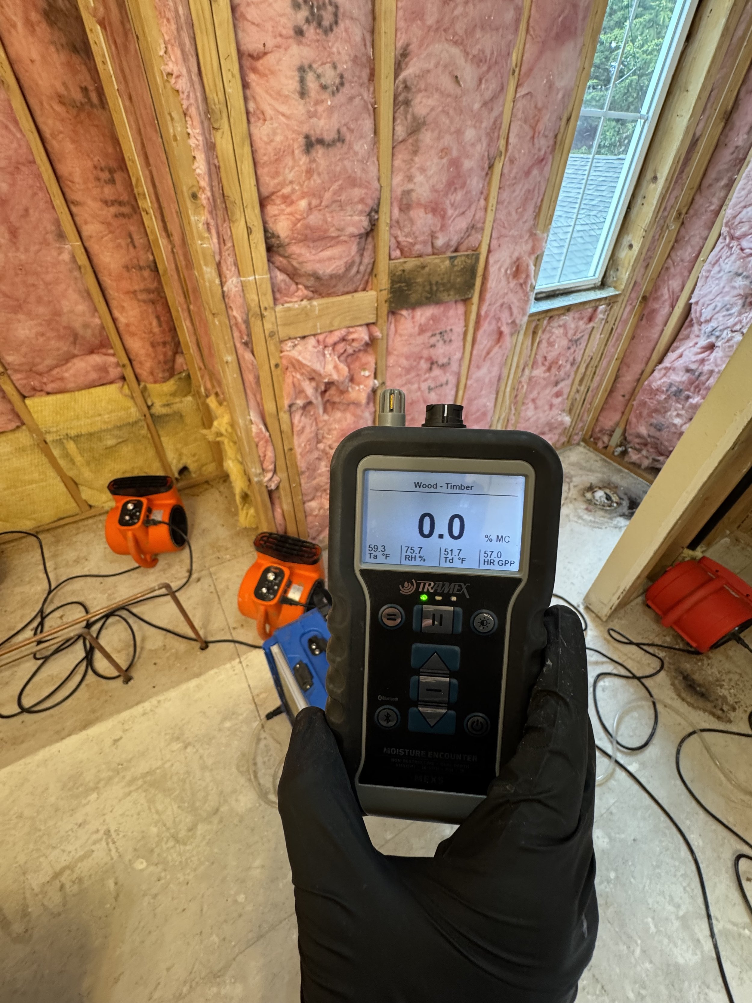 A moisture meter reading 0.0% in an unfinished room with exposed pink insulation on the walls, a small window, and two orange thermal sensors on the floor.