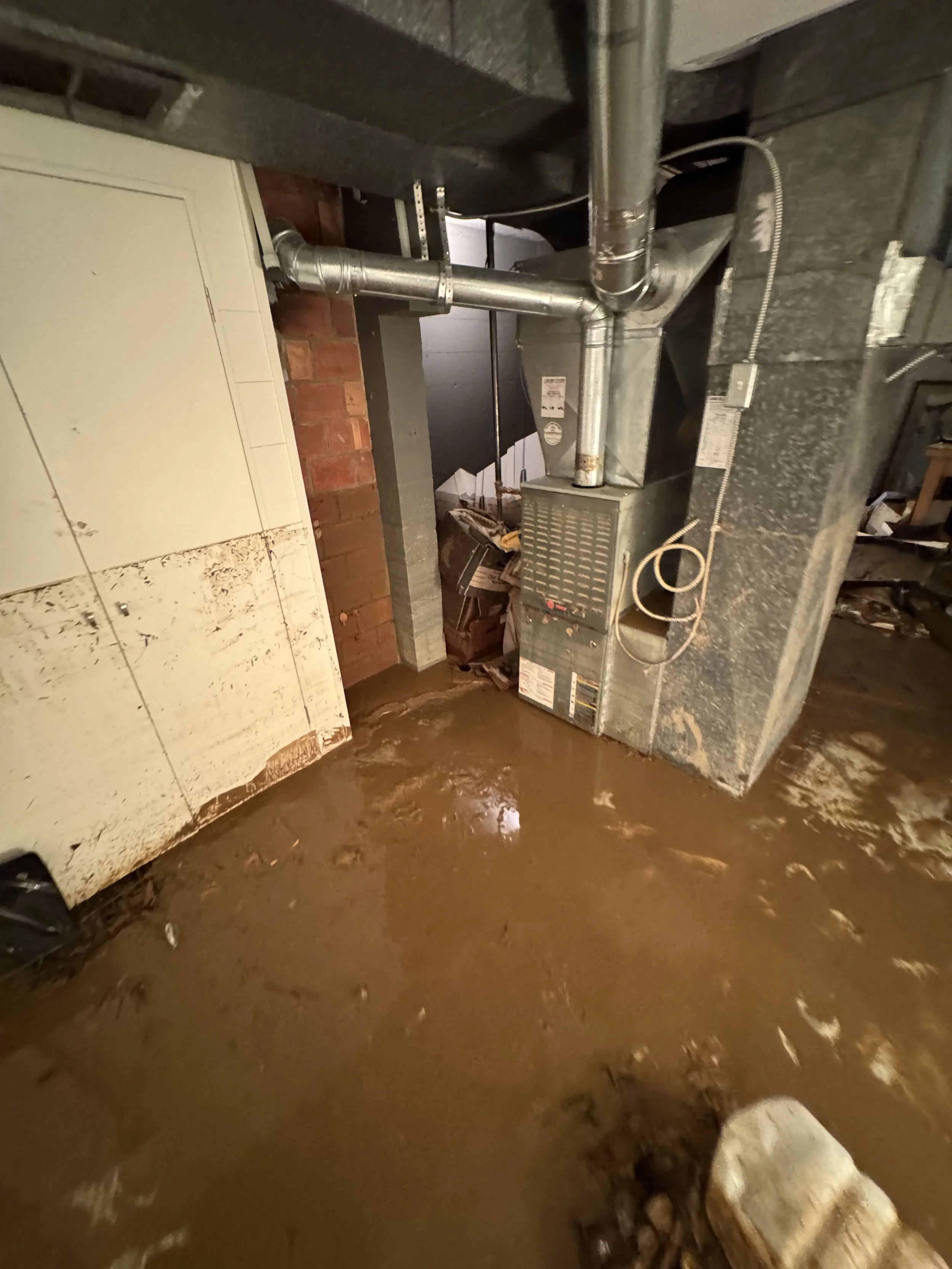 Basement with muddy floor, HVAC system, ducts, pipes, and electrical wires near the ceiling.