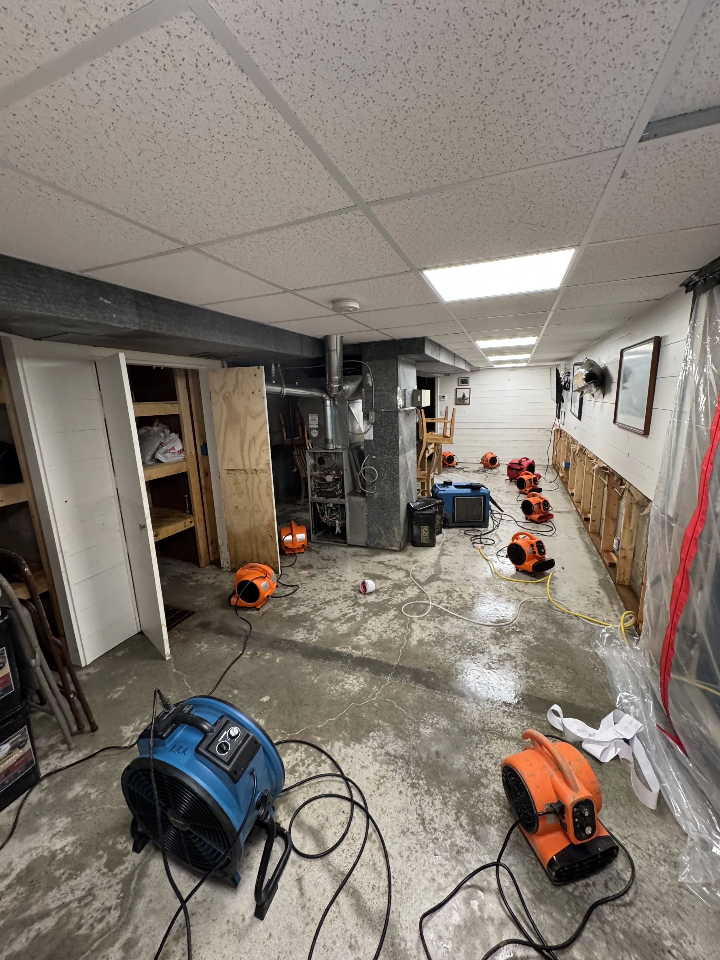 Unfinished basement with exposed wall framing, concrete floor, and installed ceiling tiles. Several portable heaters and fans are scattered throughout the space, along with some tools and equipment.