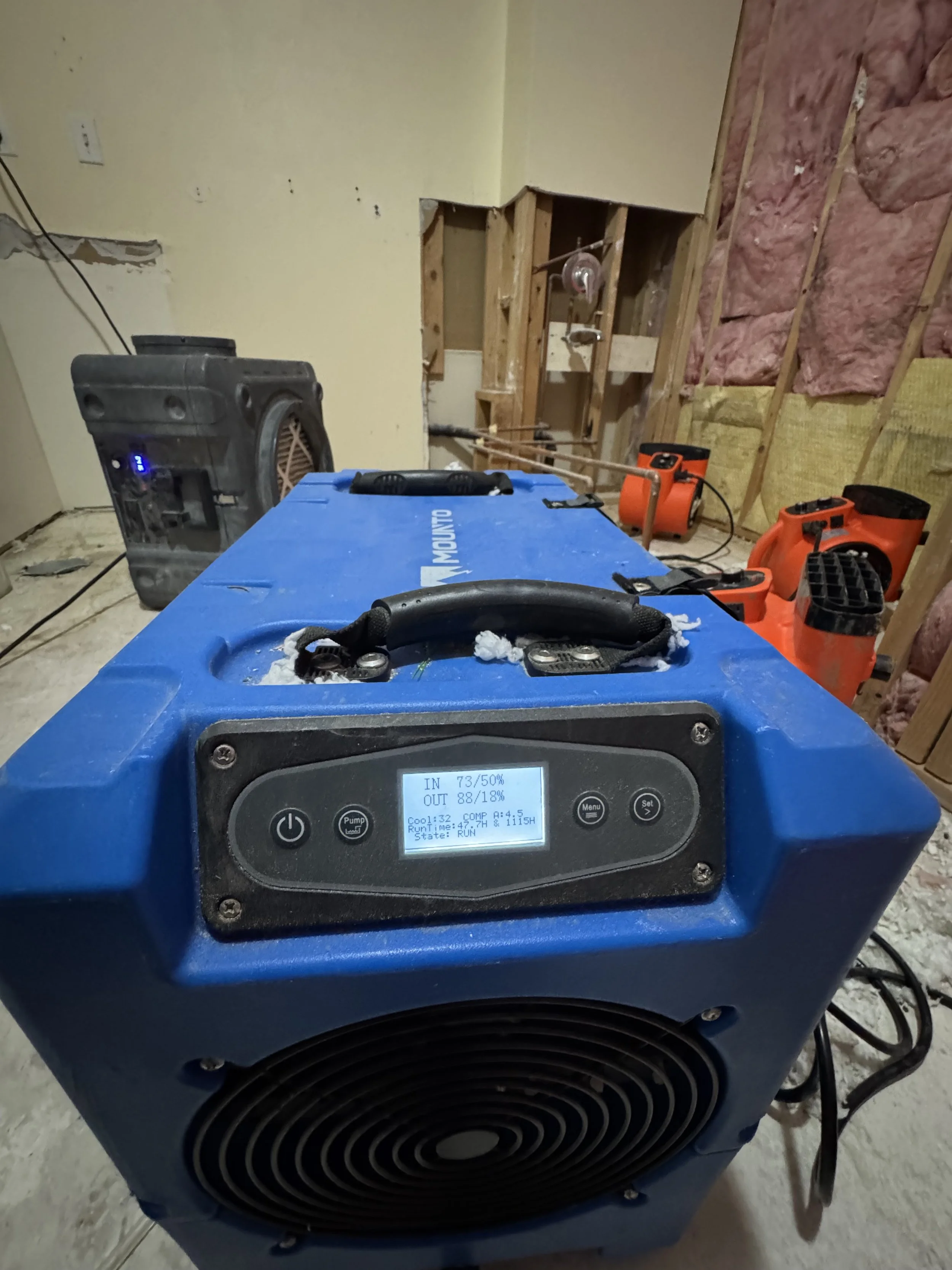 Construction site with blue portable heater on floor, surrounded by orange air movers, unfinished walls with pink insulation, and exposed wooden studs.