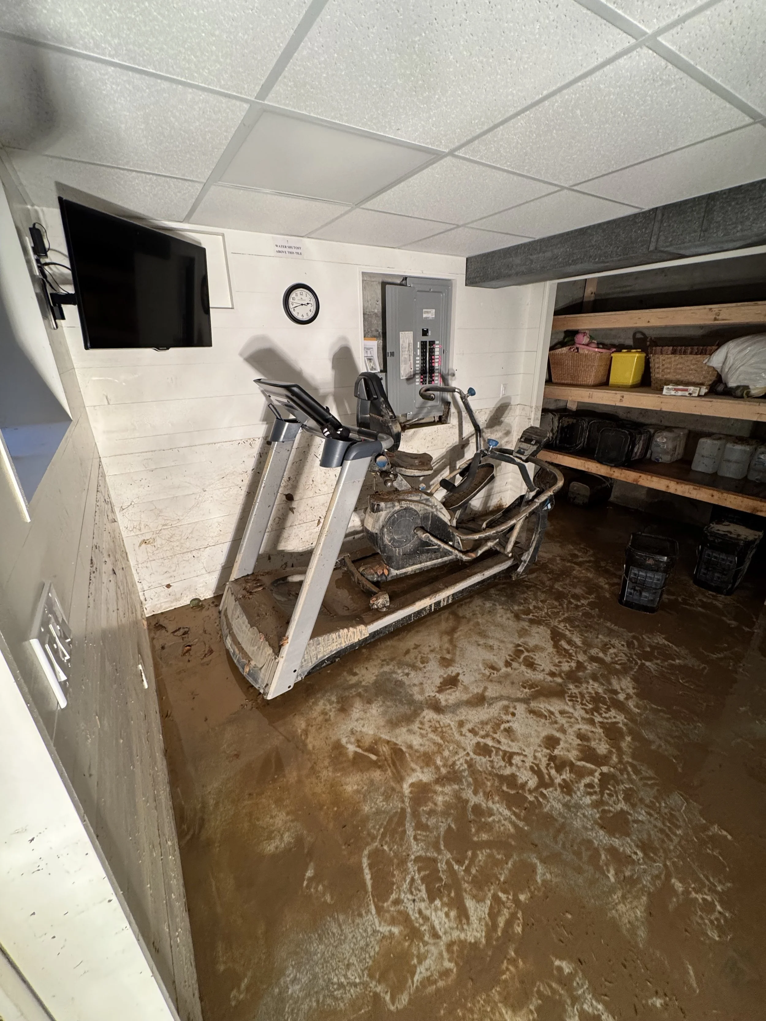 A muddy basement with an exercise treadmill covered in mud, an old damaged rowing machine, a wall-mounted TV, a clock, and a storage shelf with various items, along with flooded muddy floor.