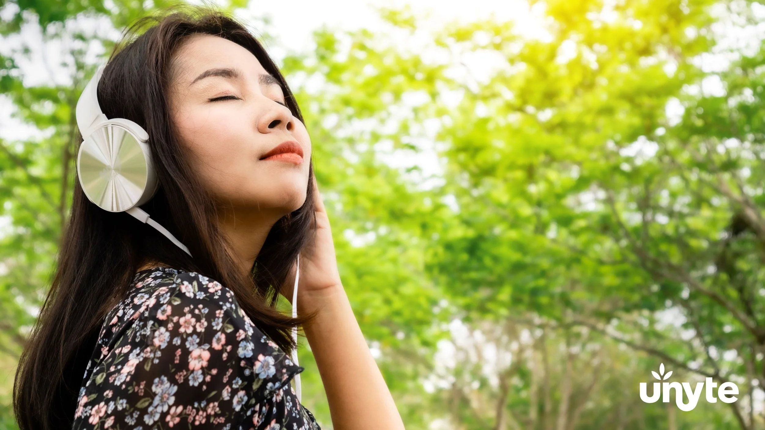 A young woman with long dark hair wearing headphones and a floral shirt enjoying the outdoors with eyes closed, surrounded by green trees and sunlight.