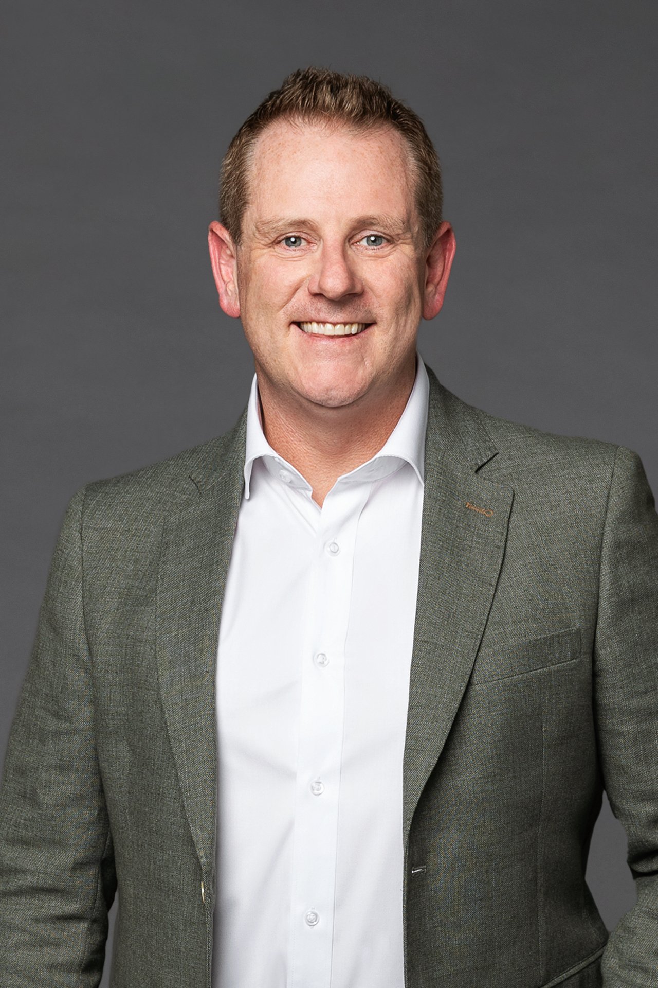 Man in a gray blazer and white dress shirt smiling at the camera against a dark gray background.
