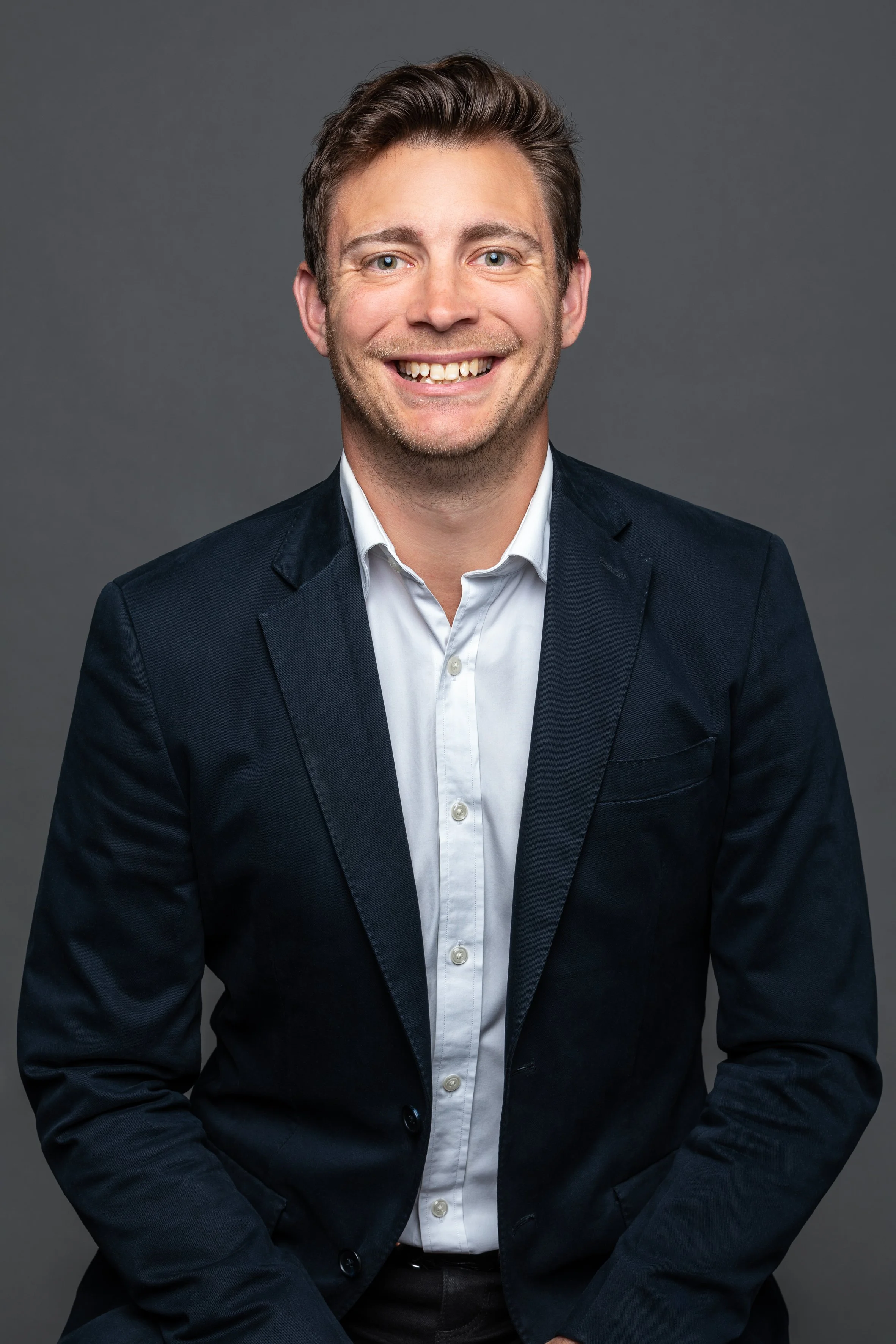 A smiling man wearing a dark blazer and white shirt, sitting against a gray background.