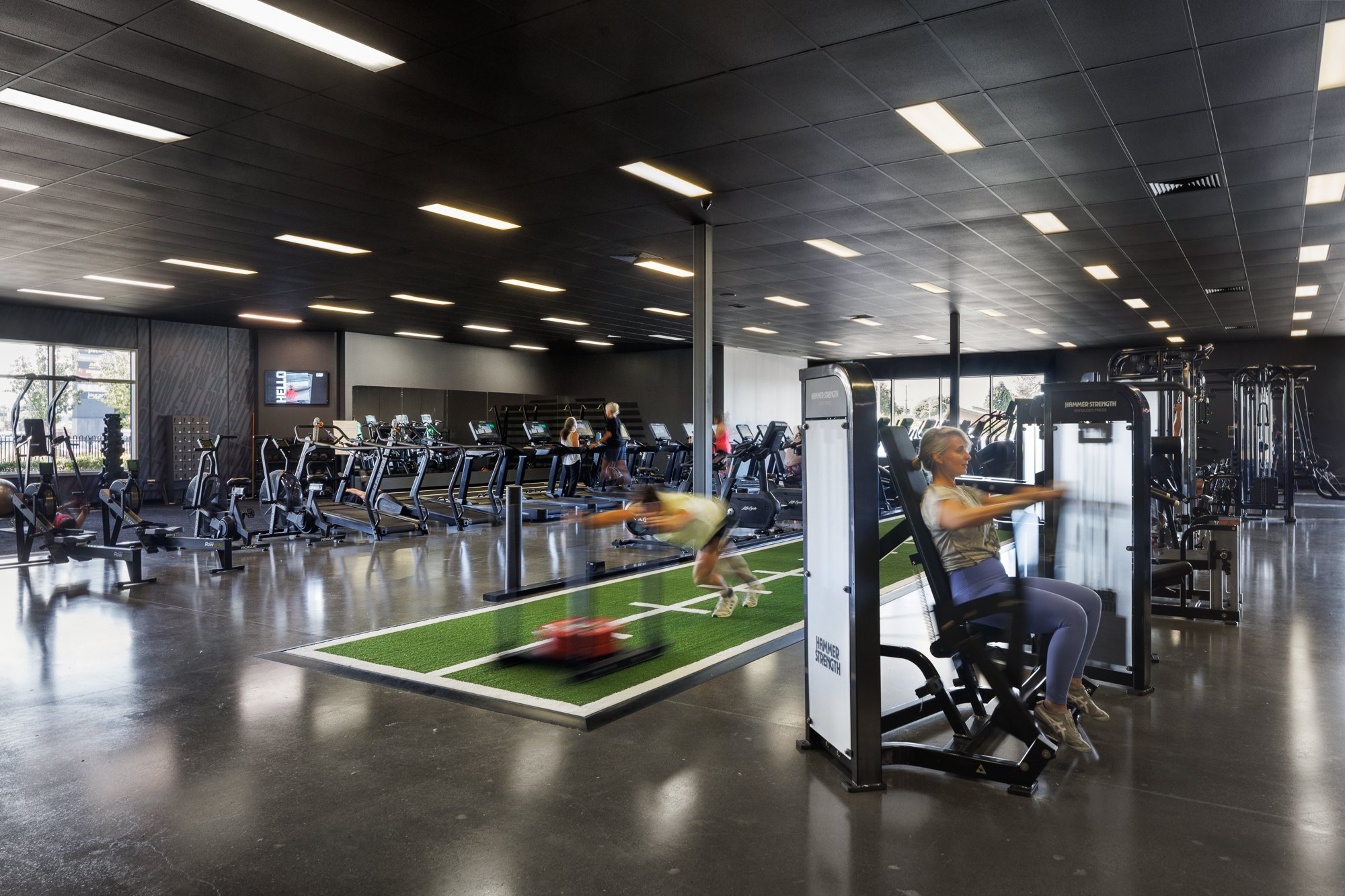 Inside a modern gym with various workout equipment, including treadmills, ellipticals, and strength training machines. People are exercising, some running on treadmills and others using strength machines, with a woman seated on a machine in the foreground. Large windows allow natural light to fill the space.