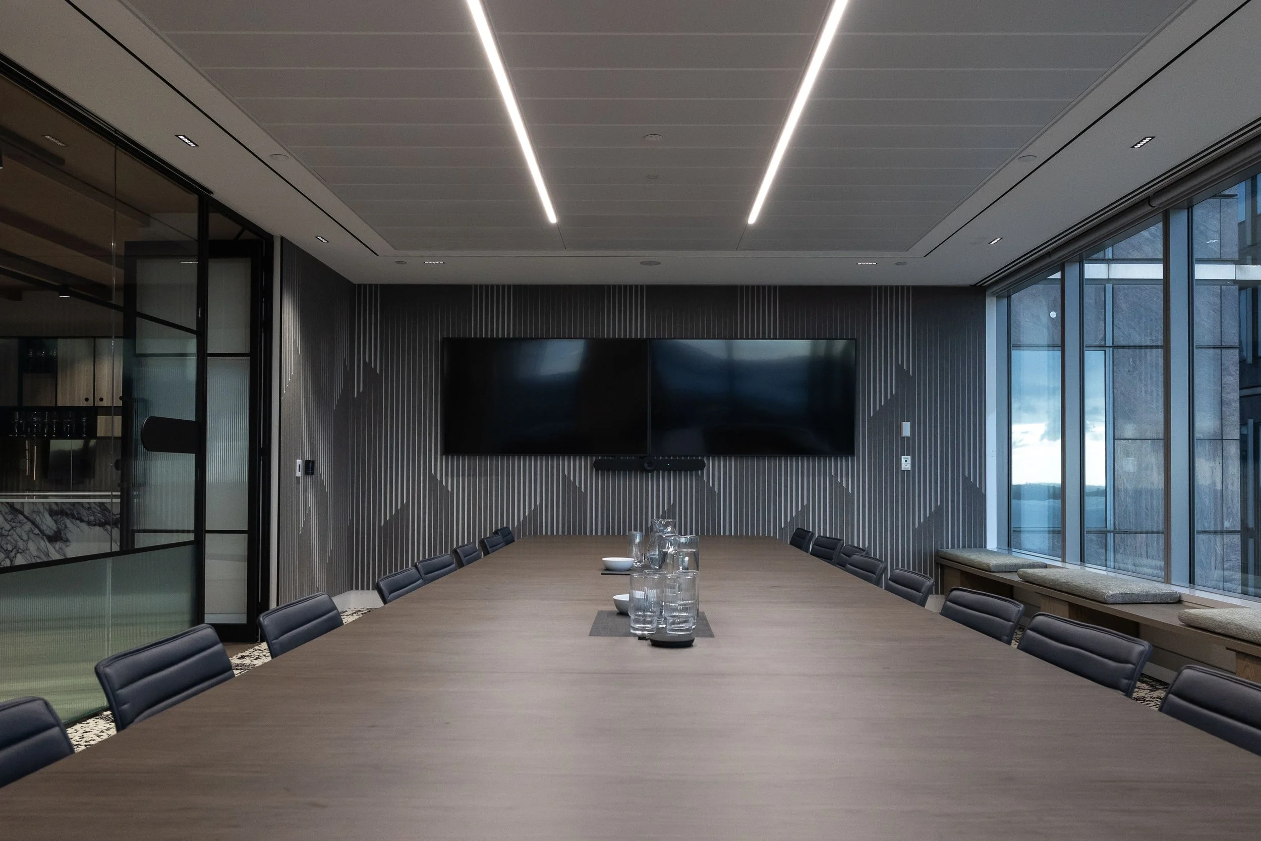 Modern conference room with a large wooden table, black chairs, a wall-mounted television screen, and windows on the right side showing an outside view.