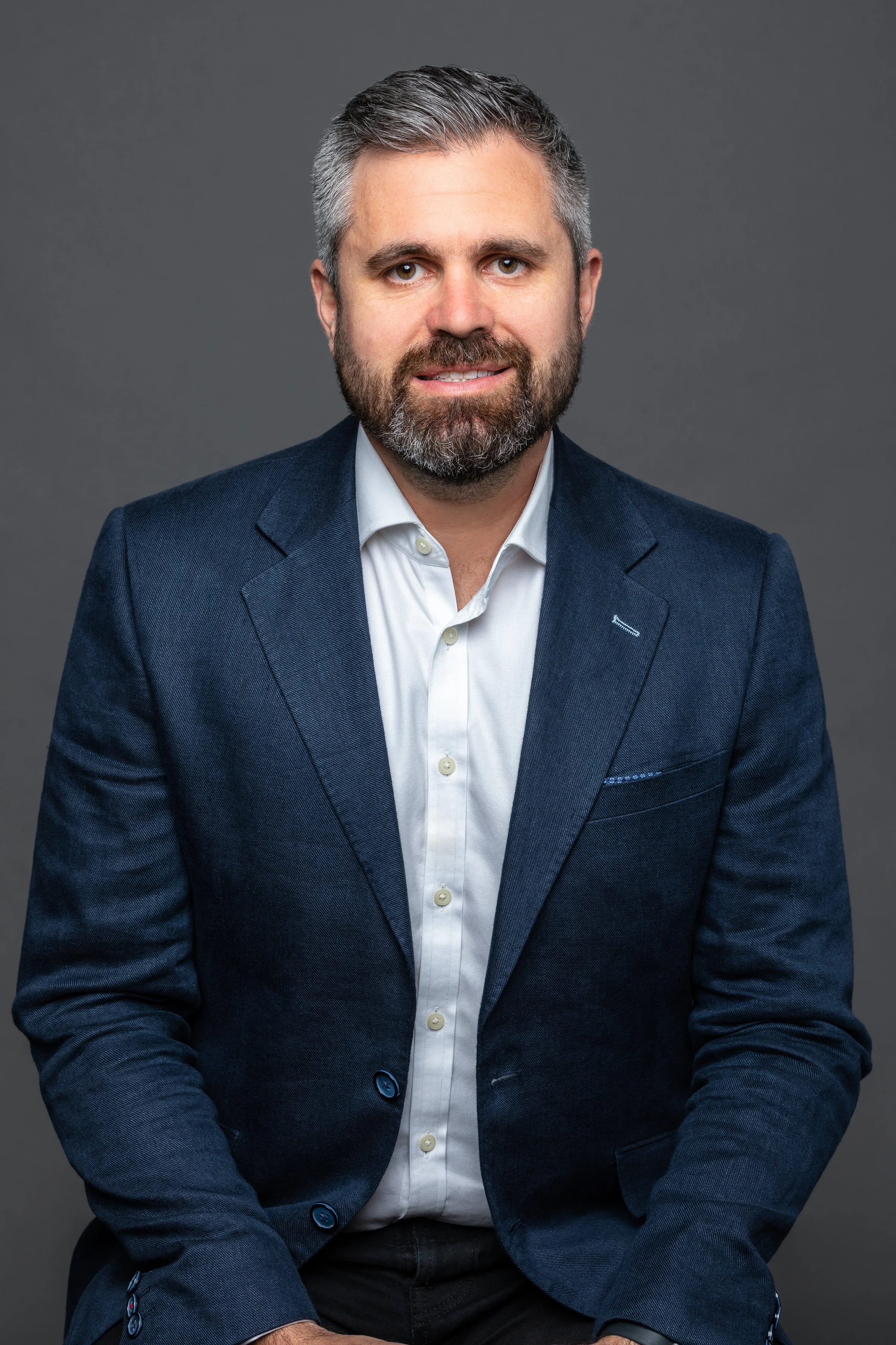 A man with gray hair and a beard, wearing a white shirt and navy blazer, sitting against a plain dark gray background.