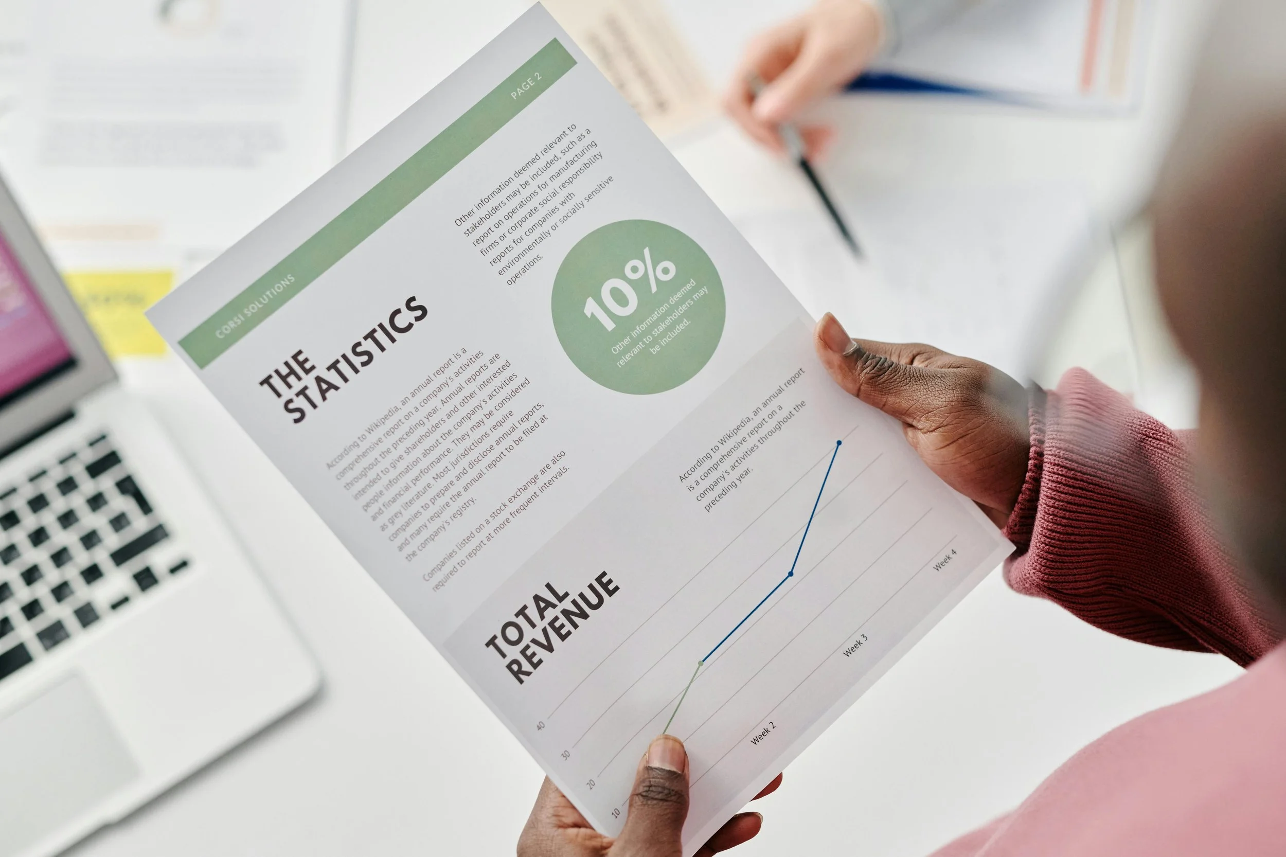 Person holding a printed report titled 'The Statistics' with a green circle showing '10%' and a line graph labeled 'Total Revenue' on a white desk, with a laptop and another person partially visible in the background.