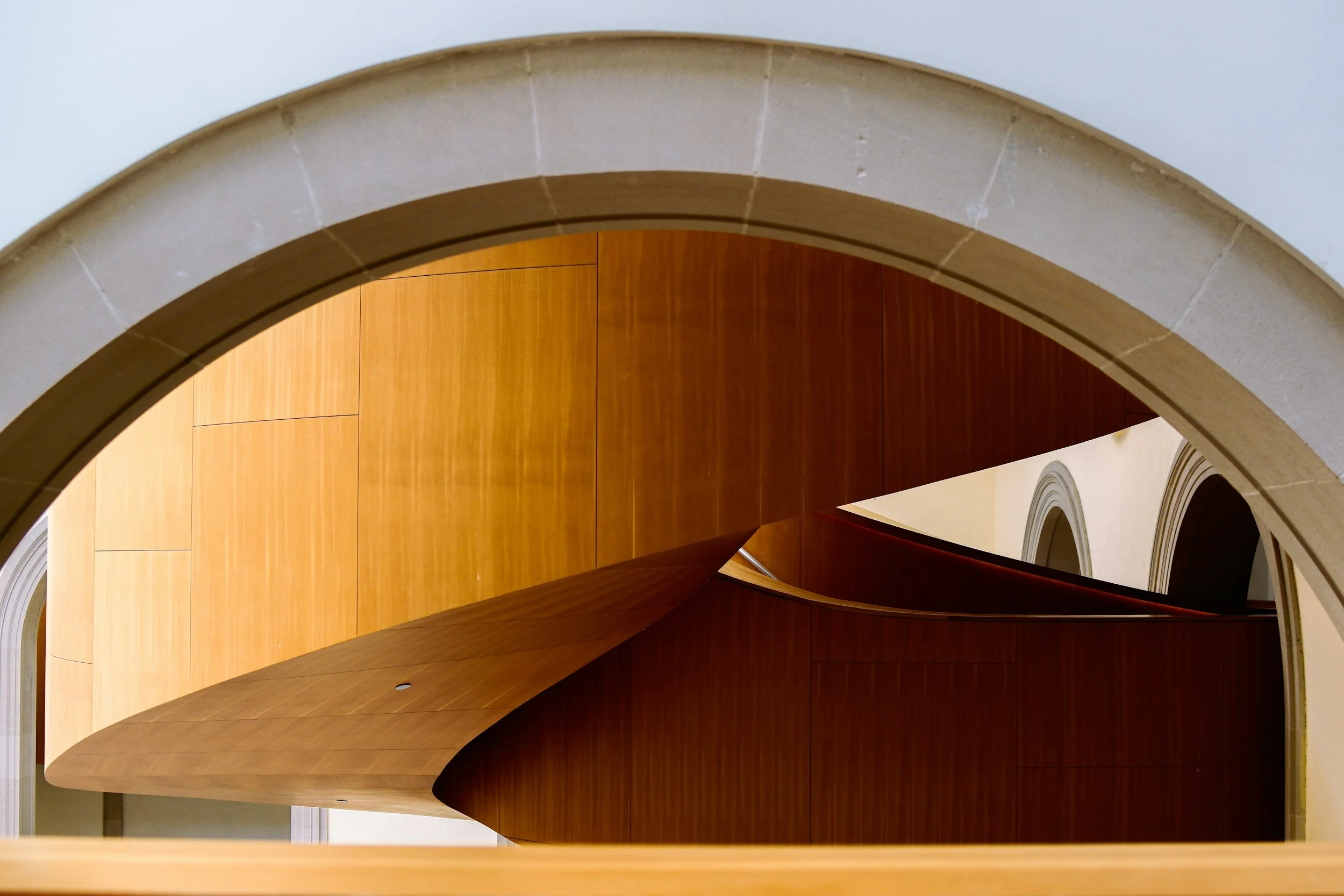 Restored curved wooden staircase framed by a stone archway, highlighting updated materials and craftsmanship within a renovated building structure.