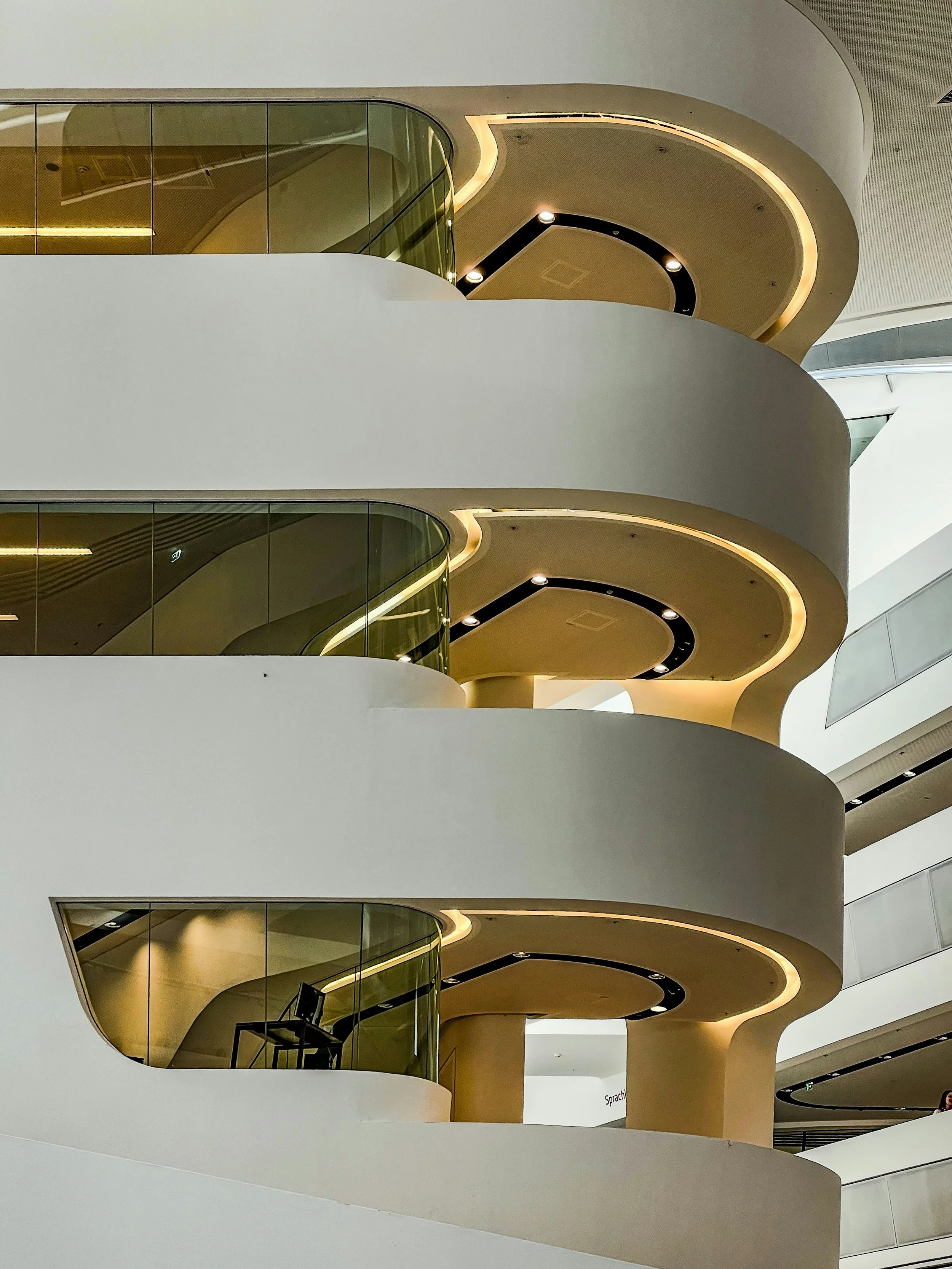 Multi-level interior view of a building with curved white balconies, glass railings, and integrated warm lighting highlighting contemporary architectural detailing.