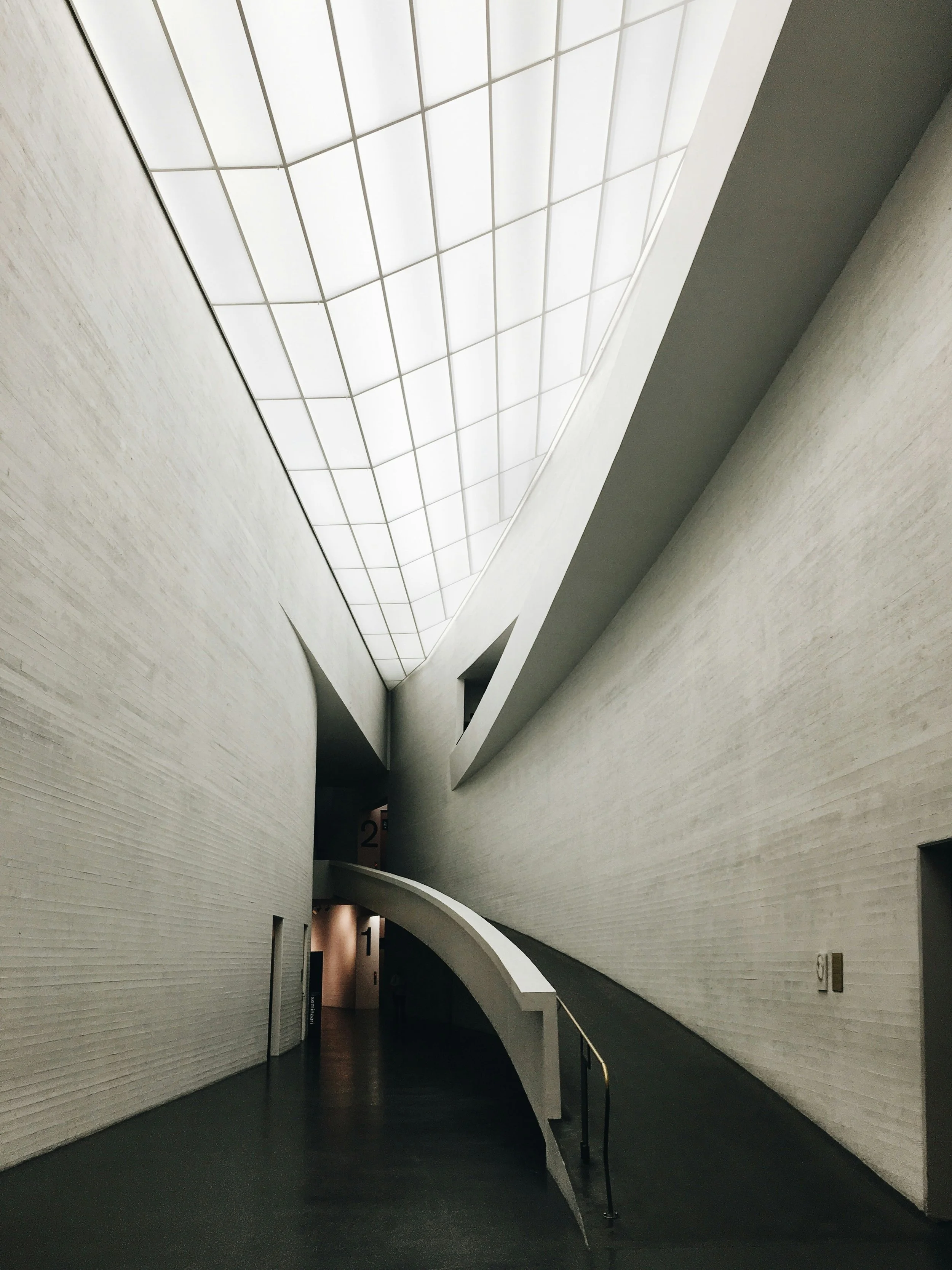 Interior corridor with sweeping curved concrete walls and a translucent grid skylight, showcasing modern minimalistic architectural design.