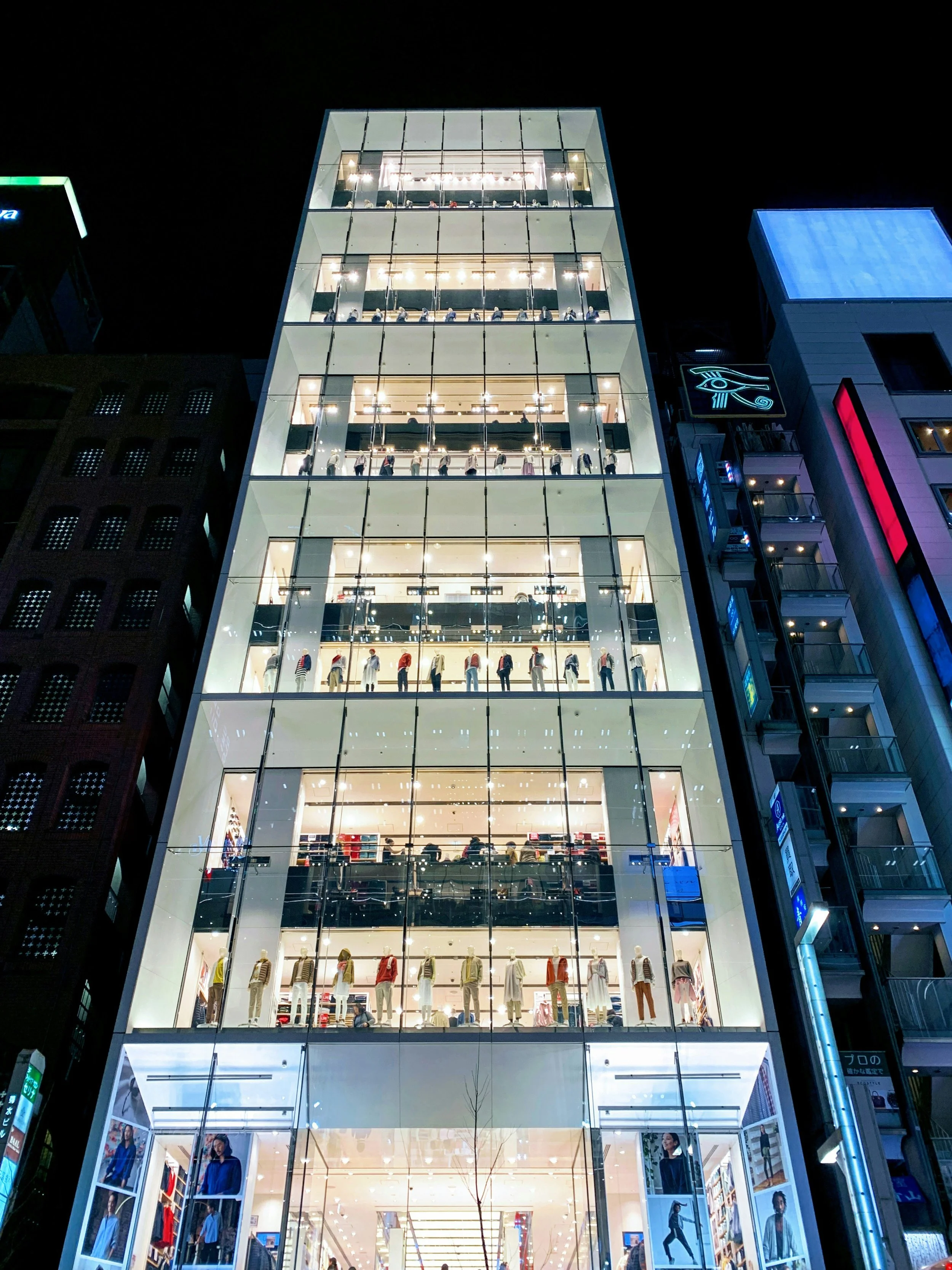 Multi-story glass building with a transparent façade showcasing exposed structural framing and open floor plates illuminated at night.