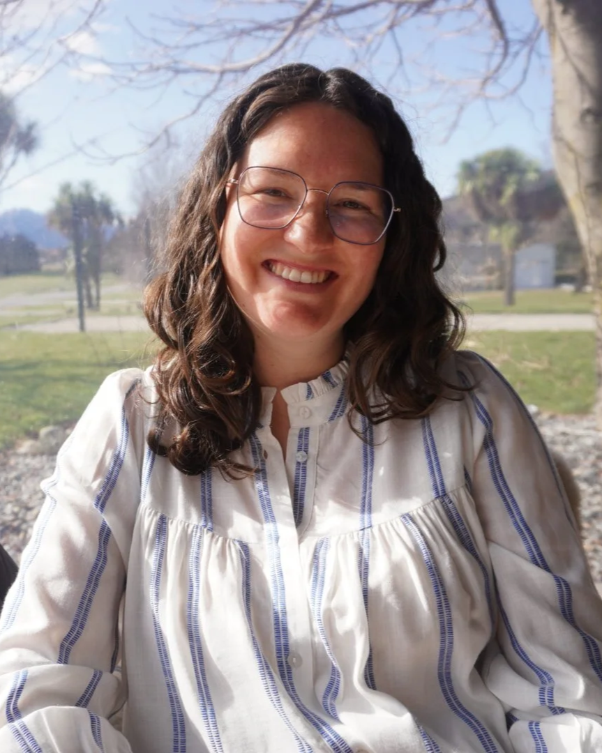 A woman with curly brown hair and glasses, smiling outdoors on a sunny day, wearing a white blouse with blue vertical stripes.
