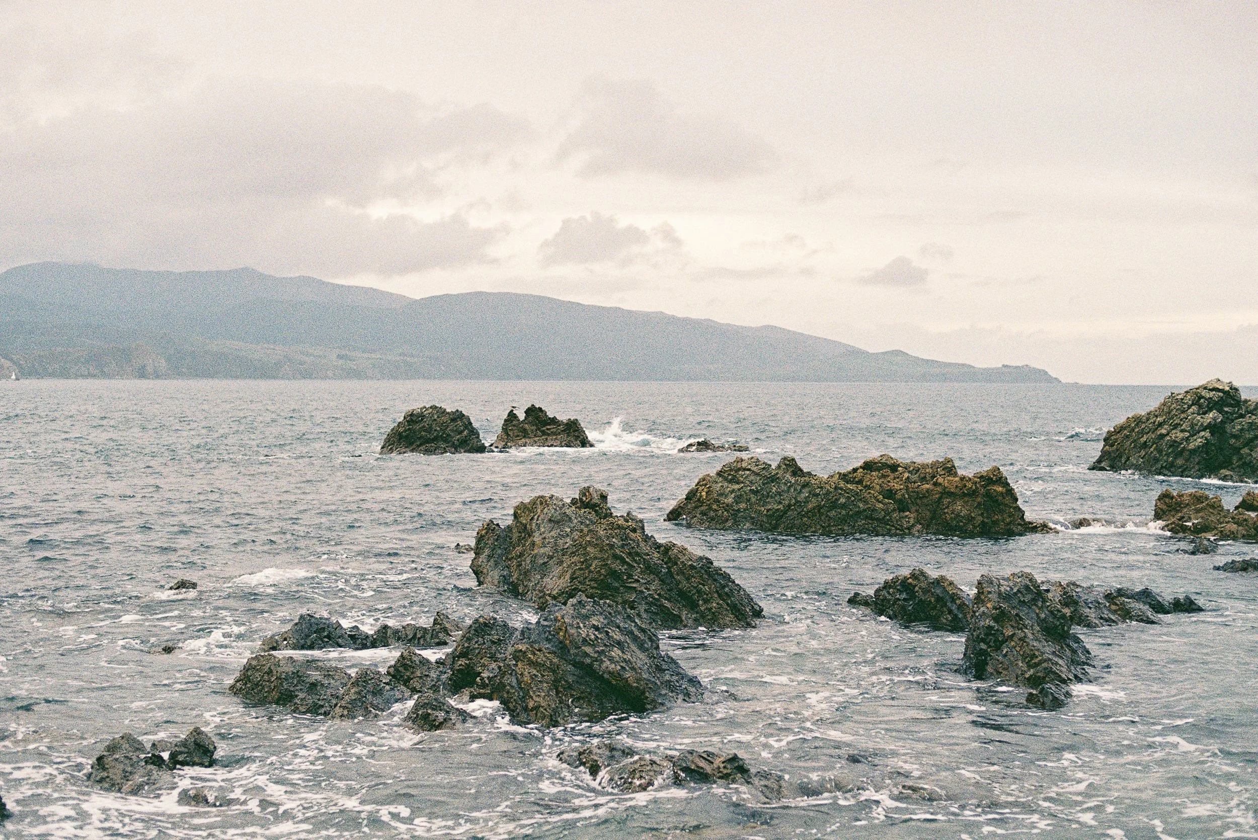 Overcast view of the ocean with rocks in the foreground and distant mountainous coastline on the horizon.