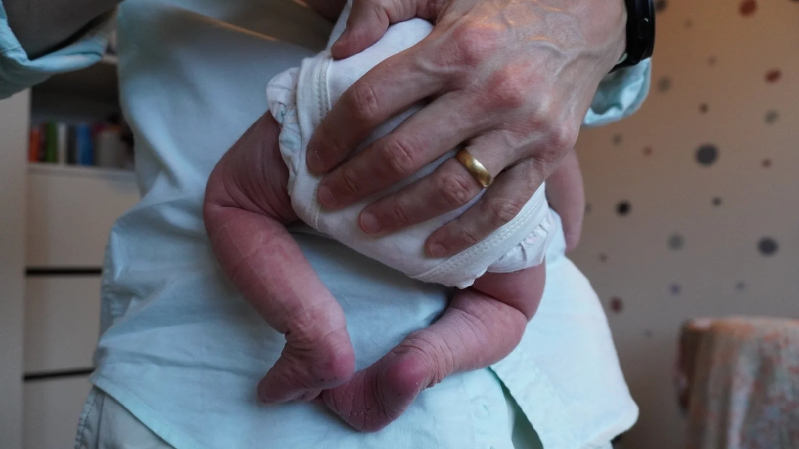 Close-up of a person holding a newborn baby, cradling the baby with both hands, one hand supporting the baby's head and the other holding the baby's body, in a hospital or medical setting.