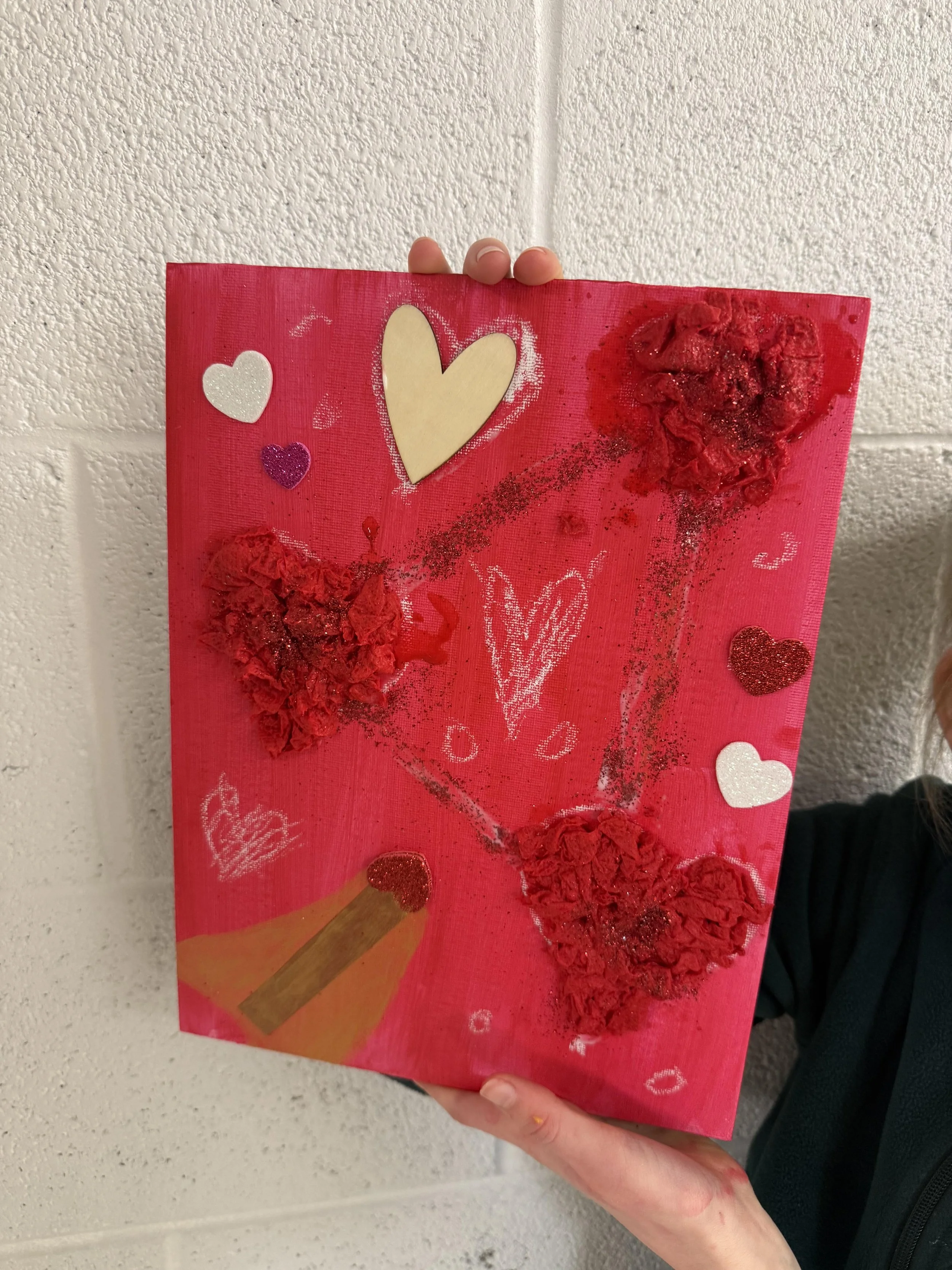 A person holding a red Valentine's Day themed poster with hearts, glitter, and textured red hearts made from crumpled tissue paper, along with white and purple heart stickers, and a crayon drawing of a heart with a name inside.
