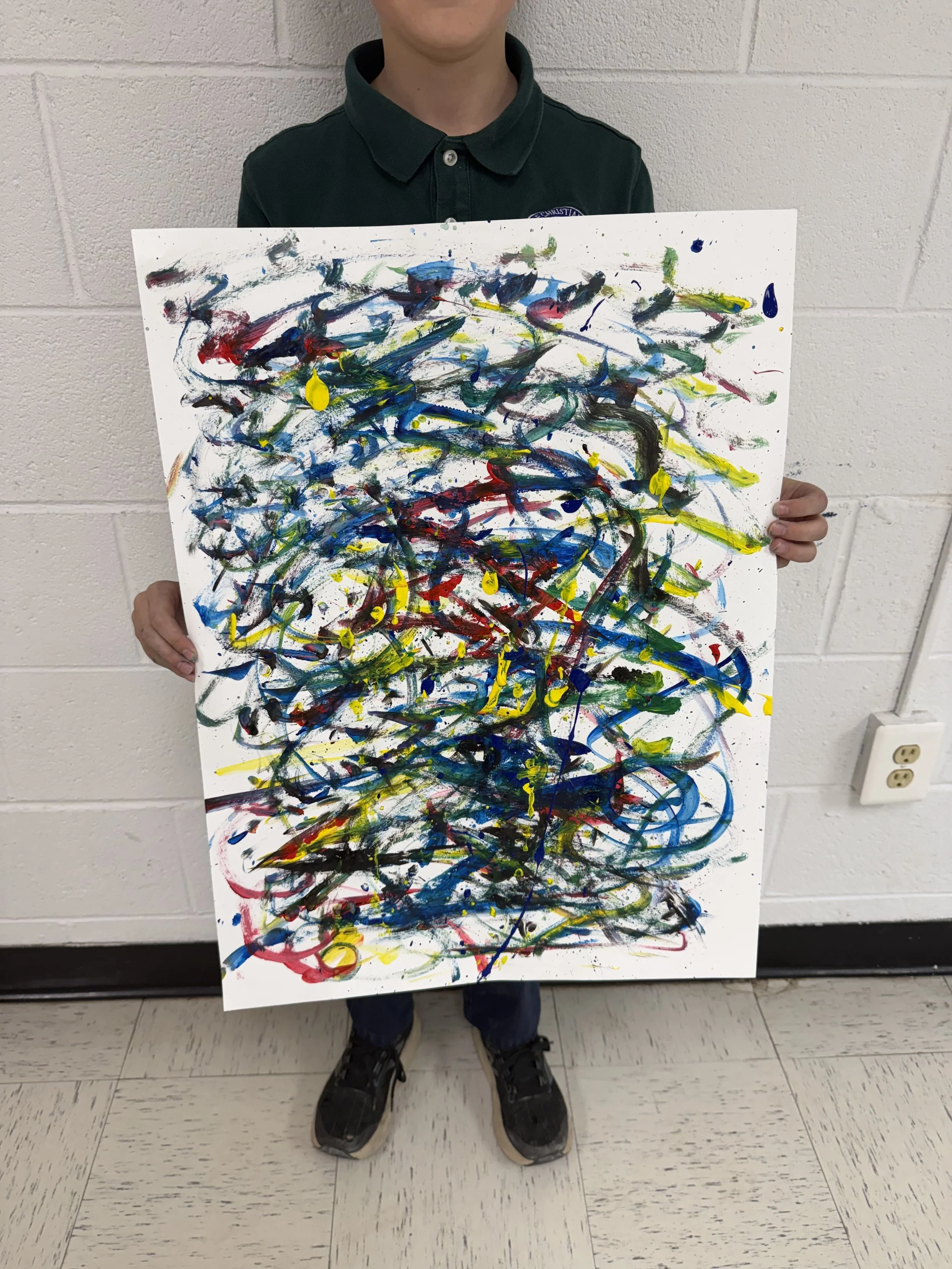 Child holding a colorful abstract painting with splashes of red, yellow, blue, green, and black, against a white wall background.