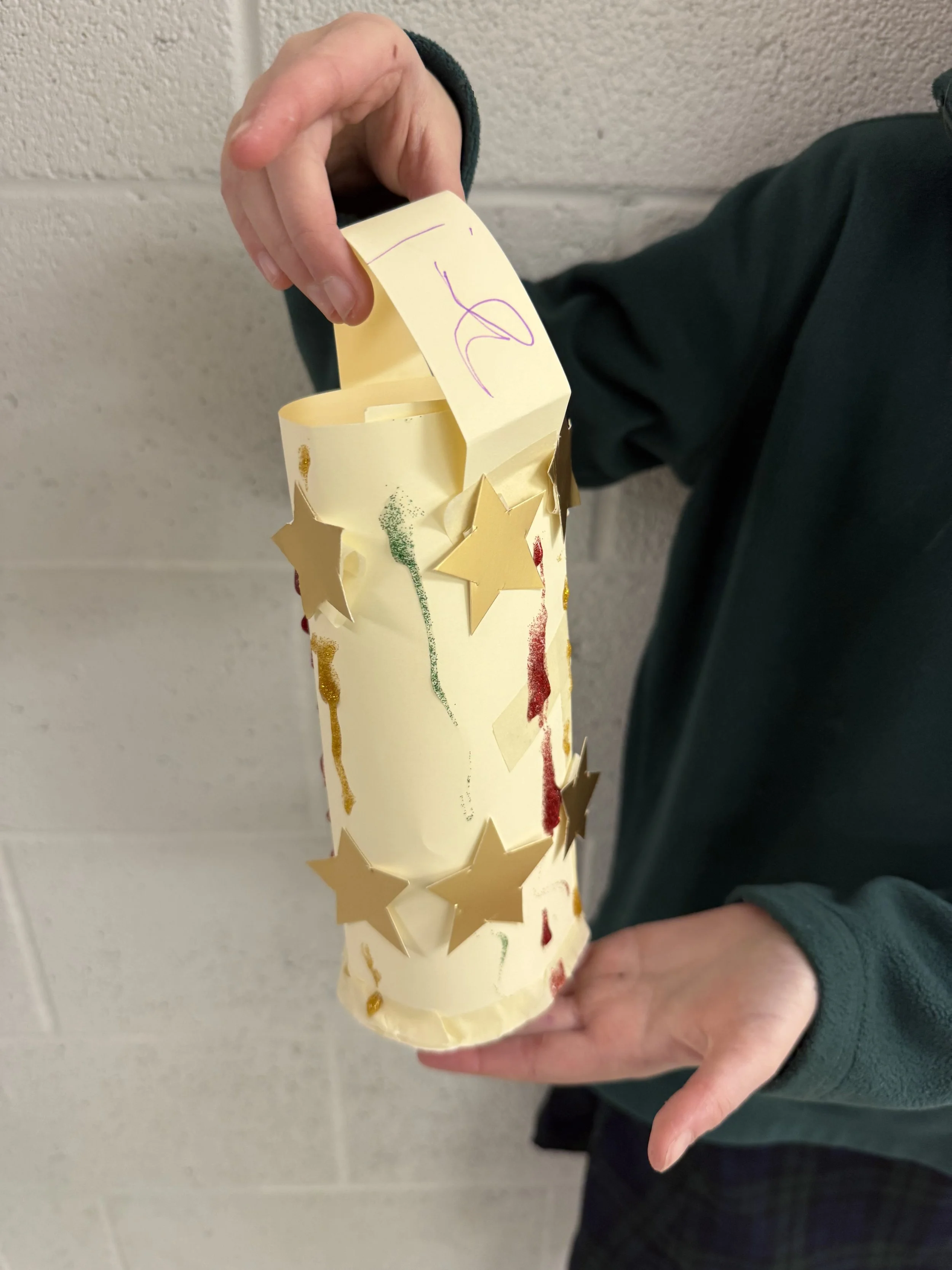 Person holding a decorated paper tube with star cutouts, glitter, and drawings, against a plain wall.