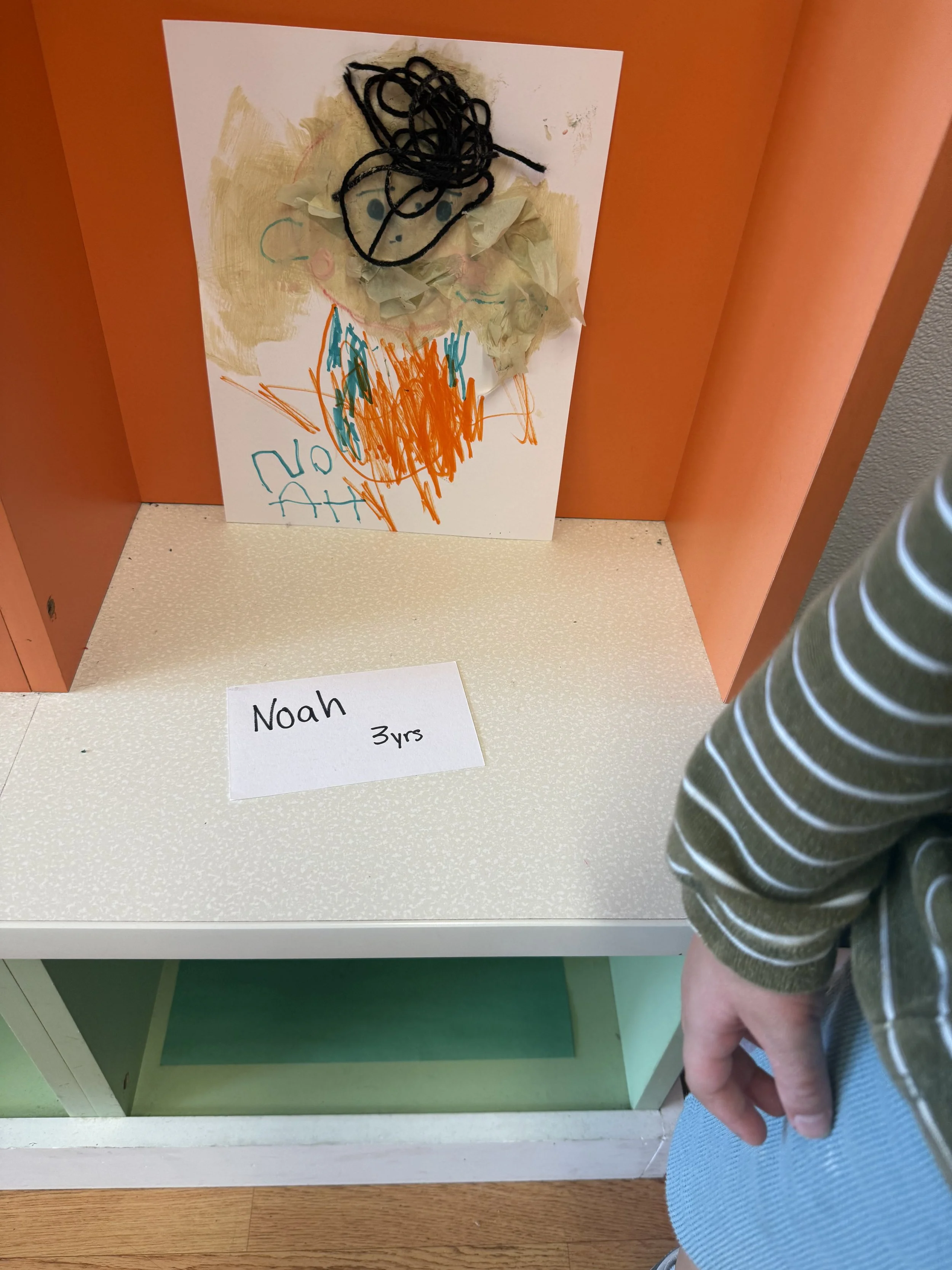 Children's art display featuring a colorful drawing with an orange and blue scribble and tissue paper attached to resemble hair. A label on the table in front reads 'Noah 3yrs'.