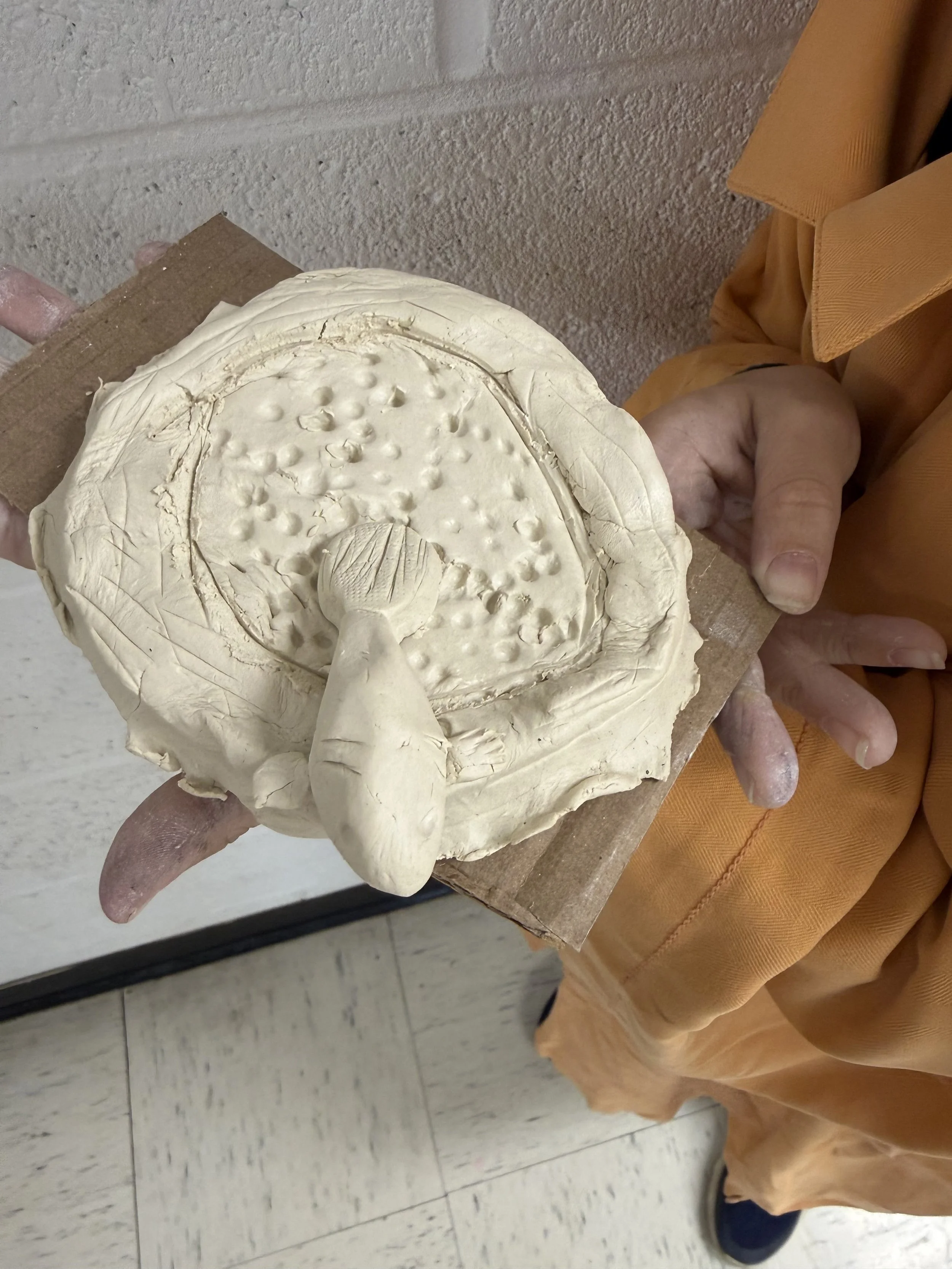 Ceramic sculpture of a mushroom growing from the center of a circular, textured surface, held by a person wearing an orange shirt.