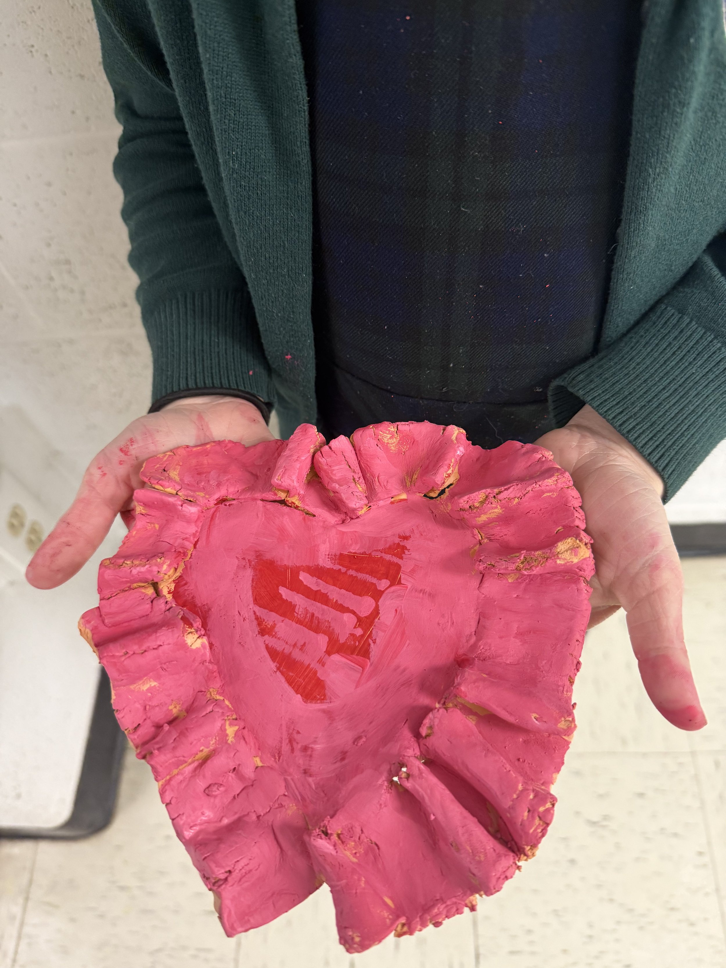 Person holding a pink, heart-shaped ceramic object with gold accents and a red heart painted on it.