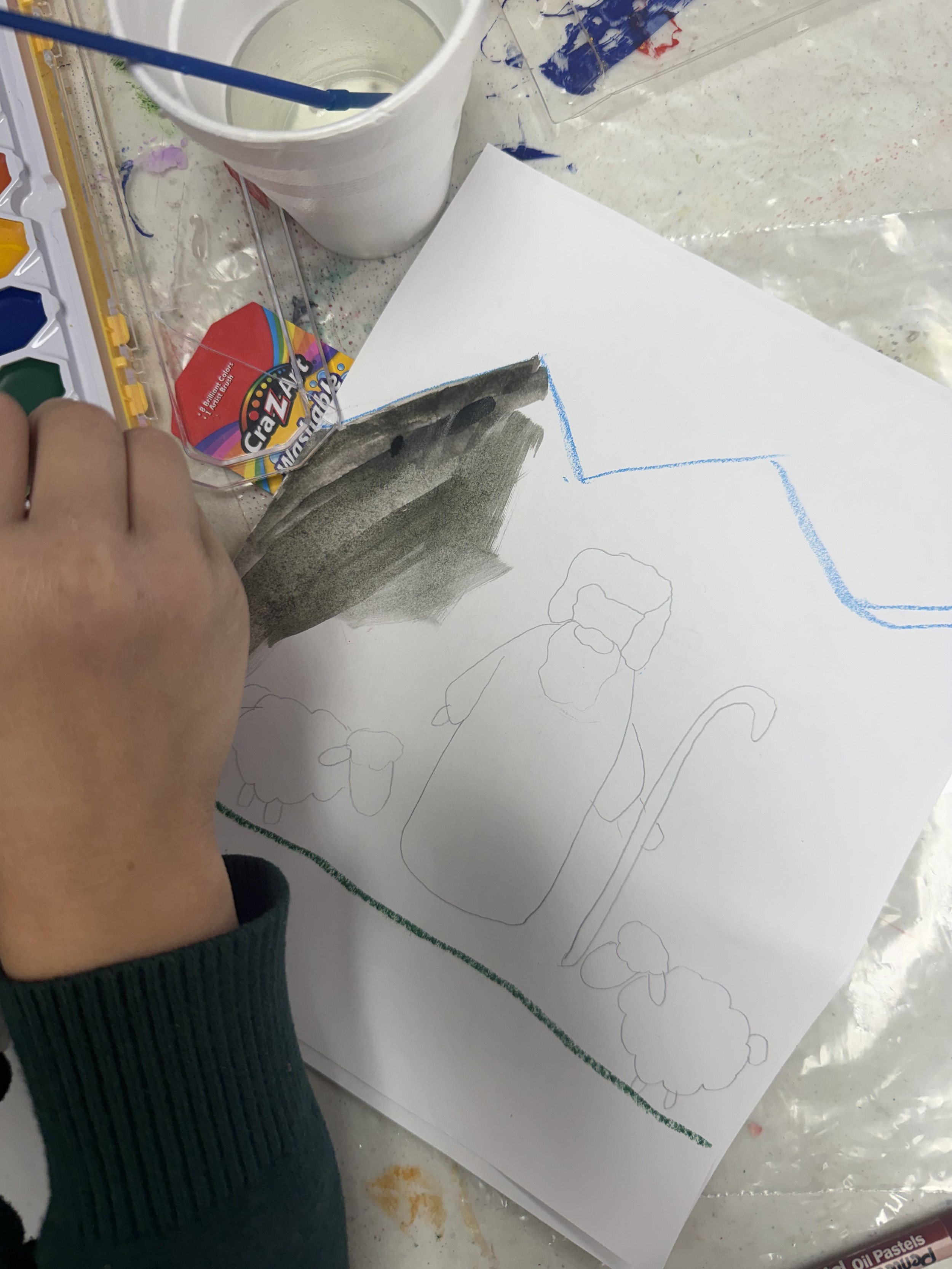 Drawing of a cat, sheep, and a bottle on paper, with art supplies and a partially colored dark shaded area nearby.