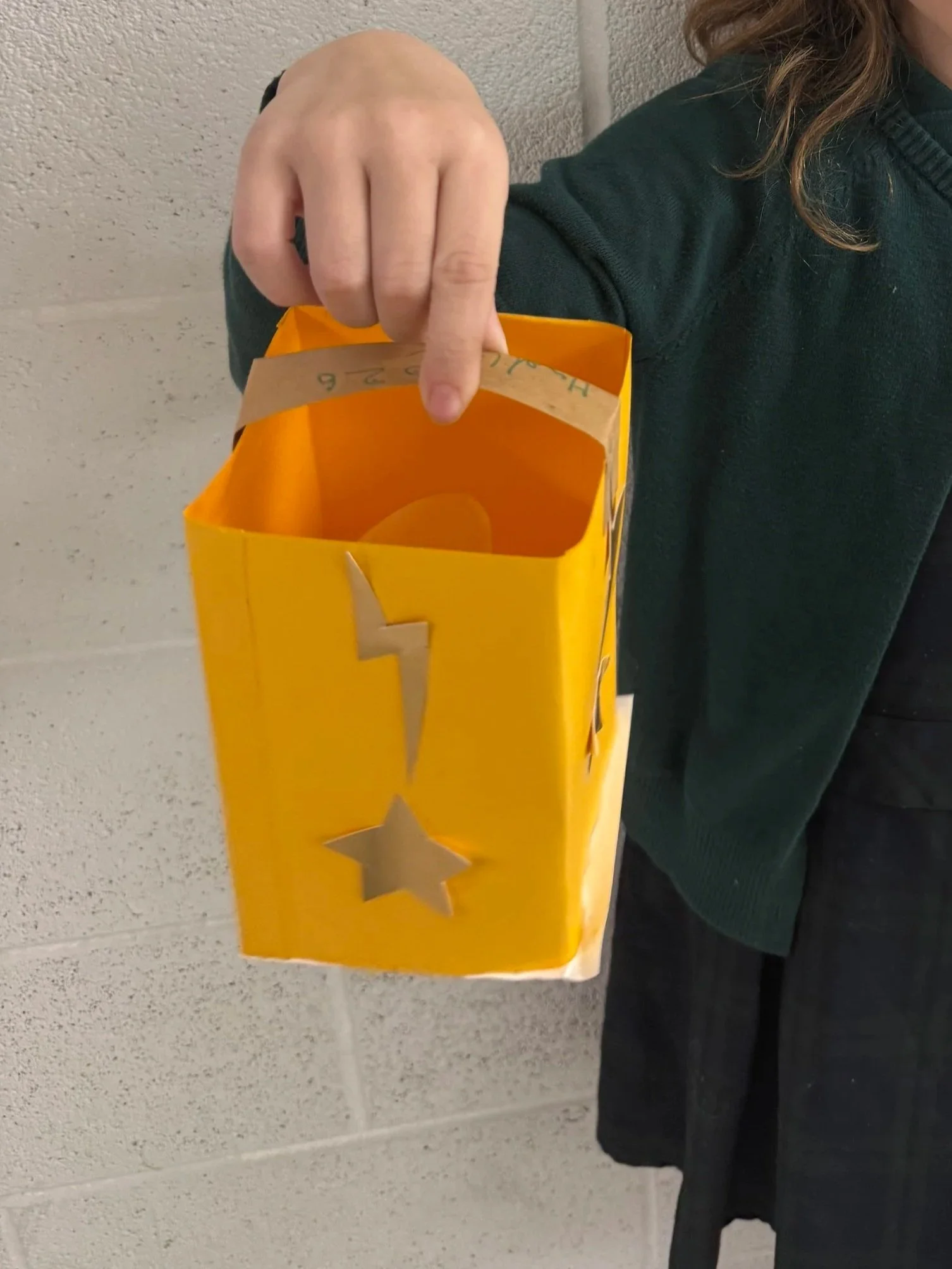Person holding a yellow paper lantern decorated with metallic star and lightning bolt cutouts, against a gray wall.