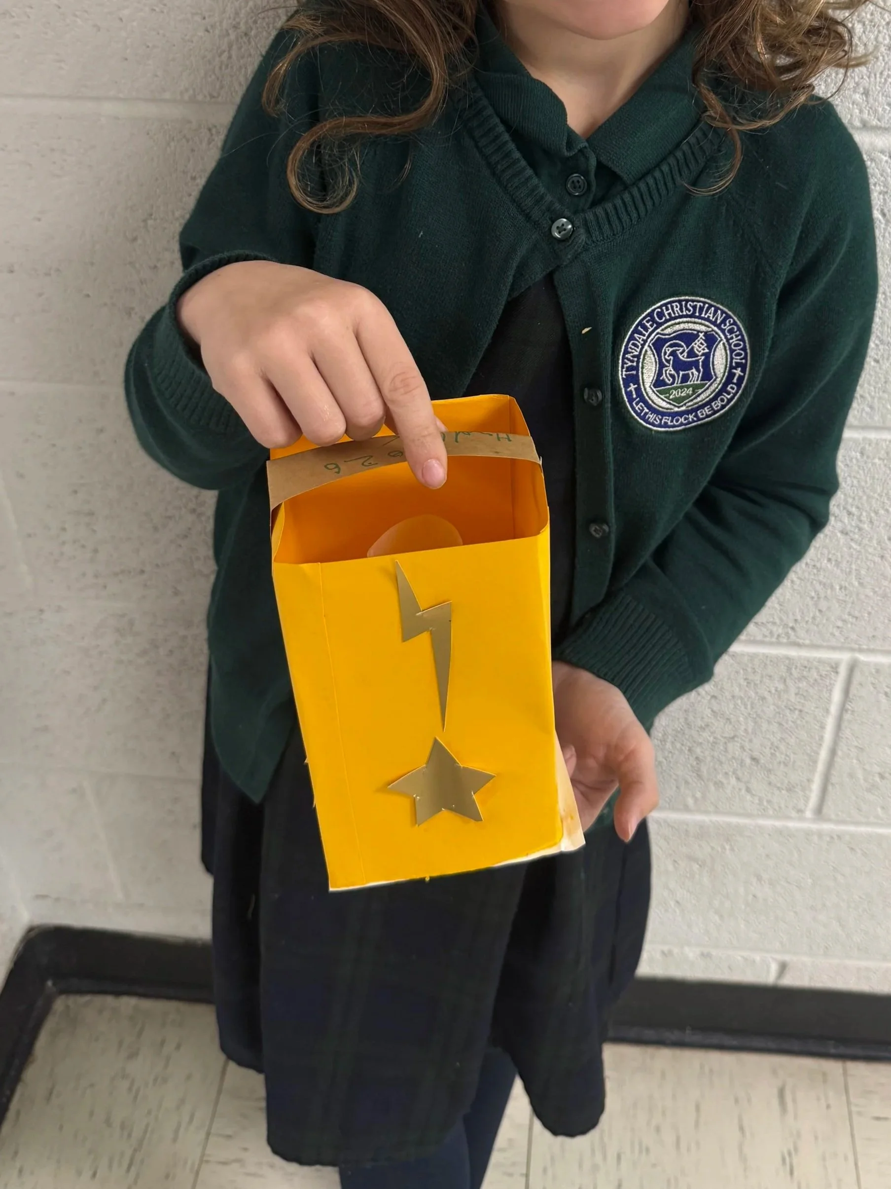 Child wearing a green school uniform holding a yellow box with silver stars and a lightning bolt cut-out.