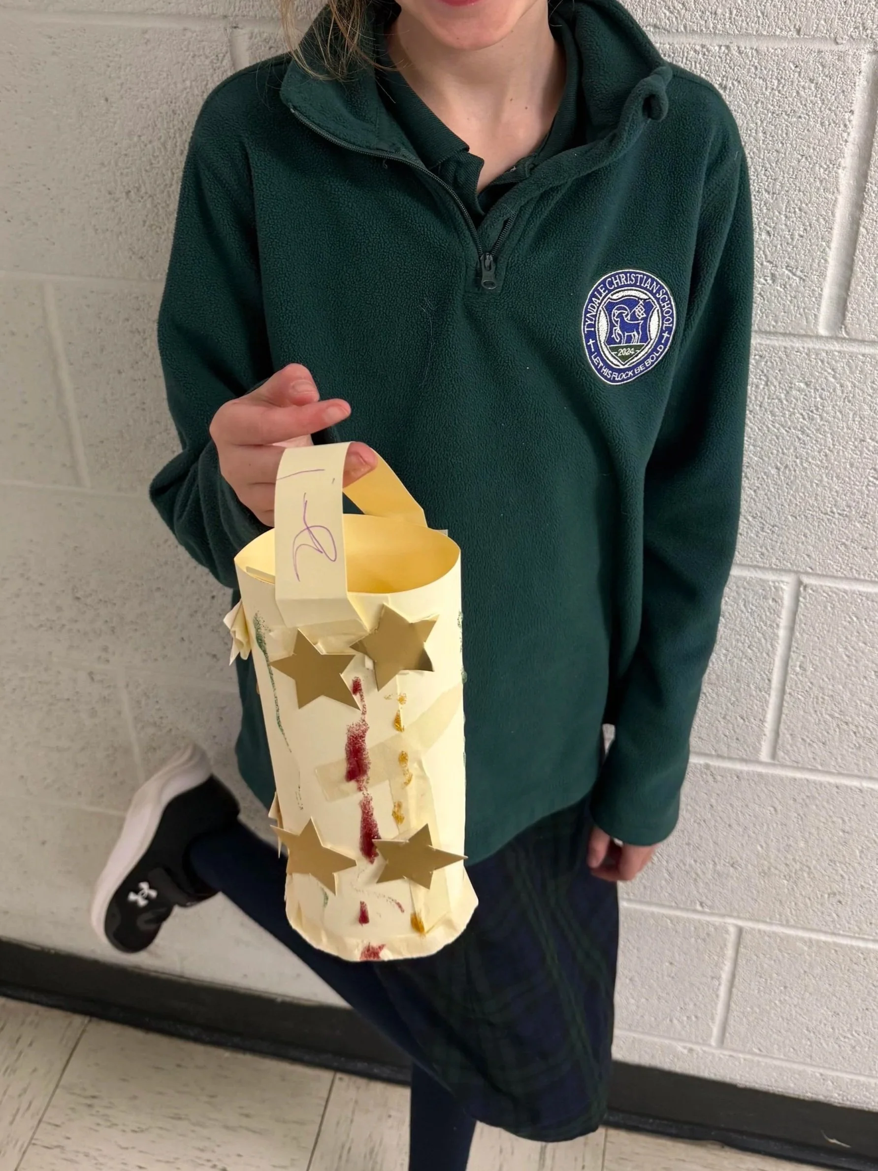 A person in a green school uniform holding a homemade lantern decorated with gold star cutouts, against a beige brick wall.