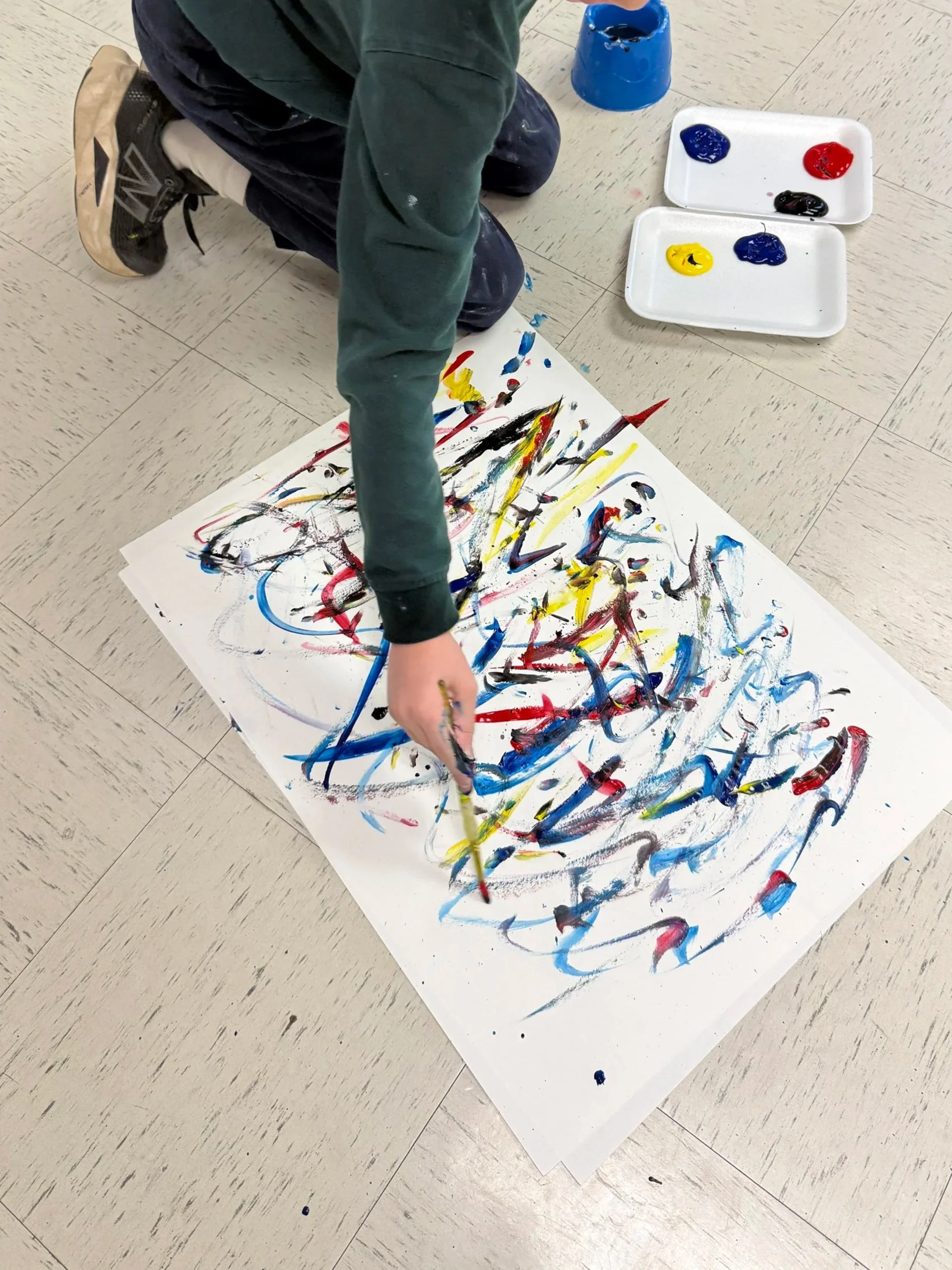 Child creating an abstract painting on large white paper with colorful paint splatters and strokes, using a paintbrush, with paint trays and a blue container nearby on a tiled floor.