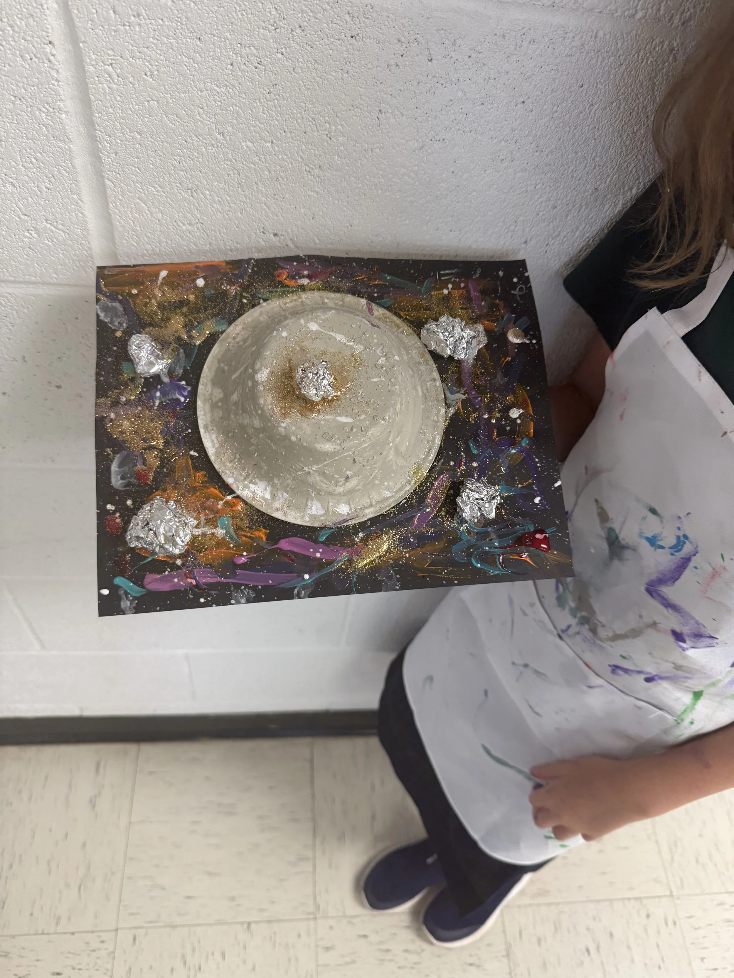 A child standing beside a painted black paper, which has a raised metallic circle in the center and small aluminum foil balls around it, representing a planetary or space-themed craft project.