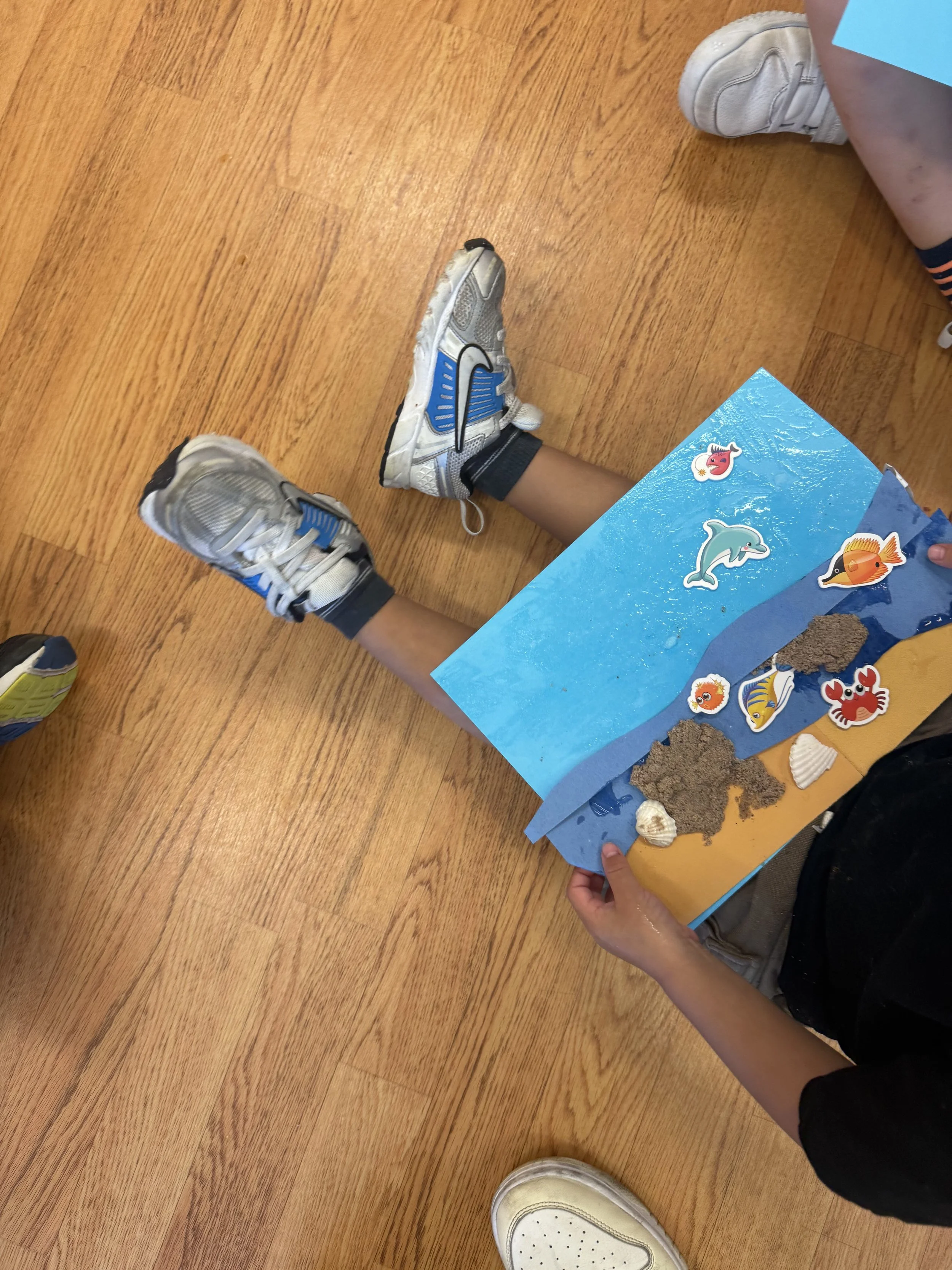 Child holding an ocean-themed craft project on blue paper, decorated with stickers of dolphins, fish, and other sea creatures, with sand and seashells glued on.