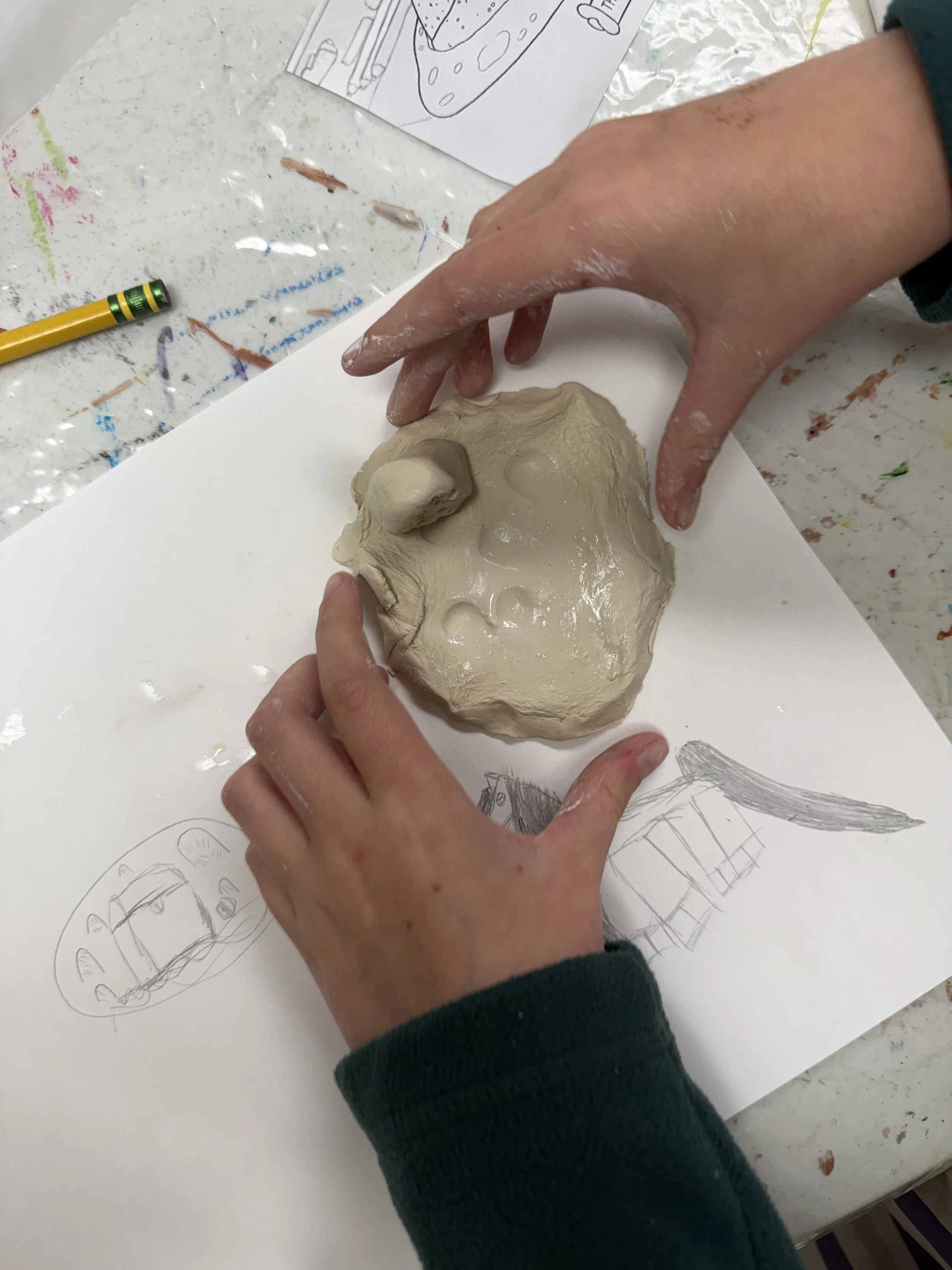 Person shaping clay into a sculpture on a white sheet of paper with sketches and drawings of objects nearby.
