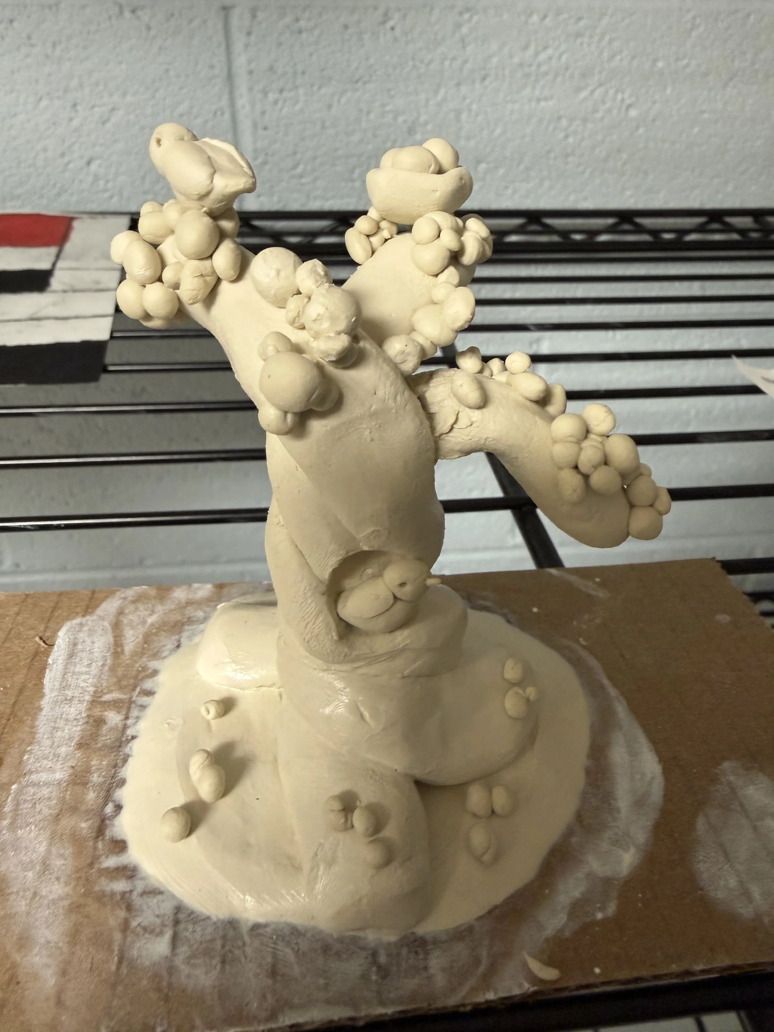 A sculpture of a gnarled, leafless tree with rounded, cork-like growths on its branches, created from a beige clay material. The sculpture sits on a square base with some scattered clay parts around it.