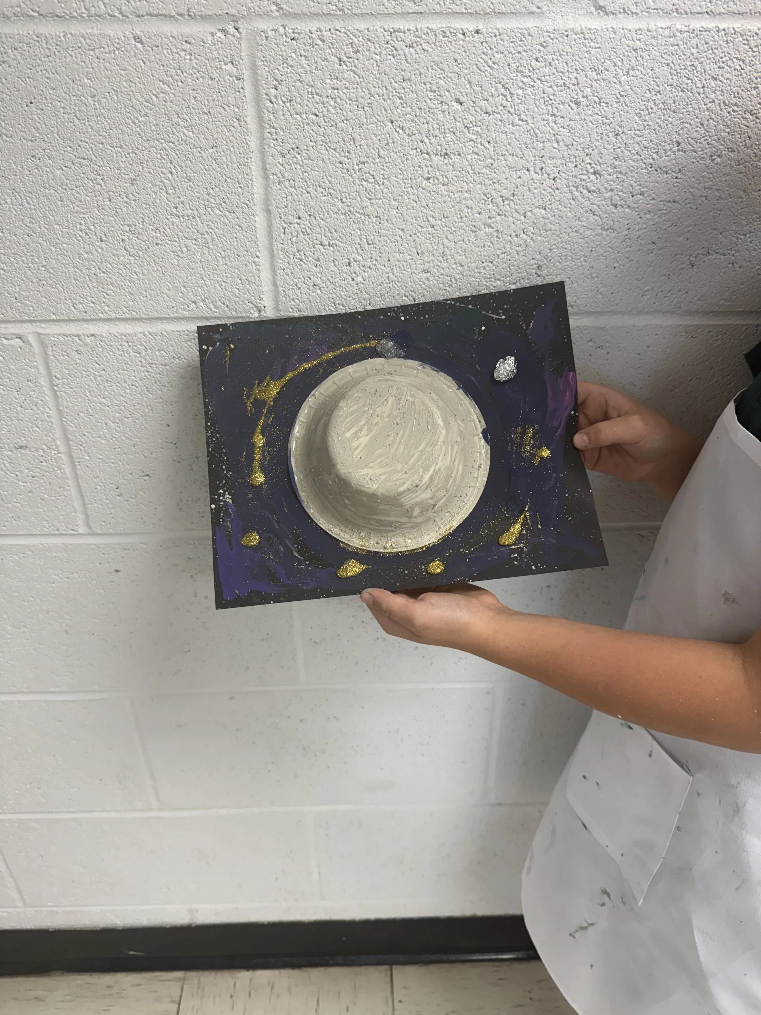 Child's hand holding a painted craft project resembling a galaxy with a moon in the center, on black paper with gold, purple, and blue splashes.