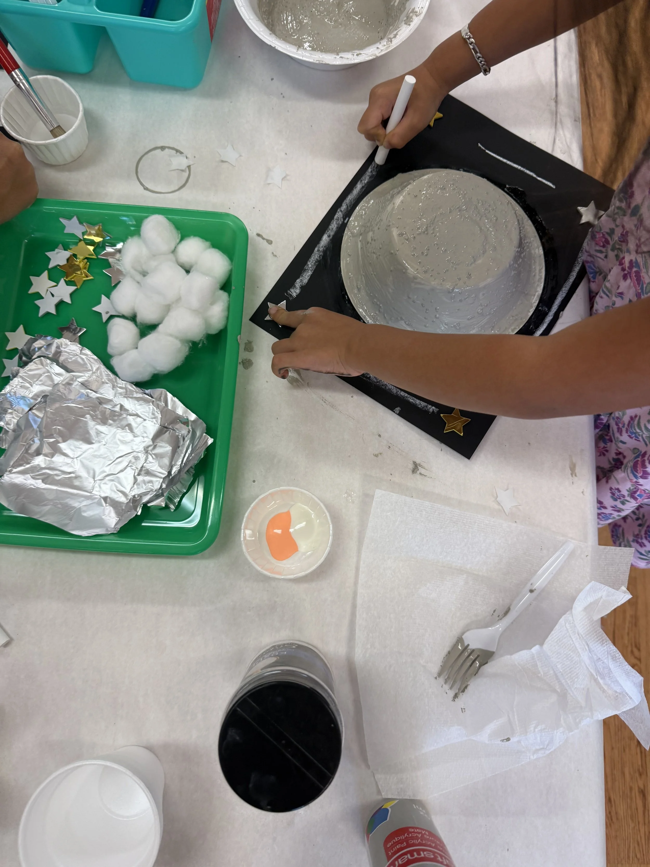 People decorating a cake with star-shaped and cotton ball decorations, using a white marker on a black board, with various decorating supplies and drinks on the table.