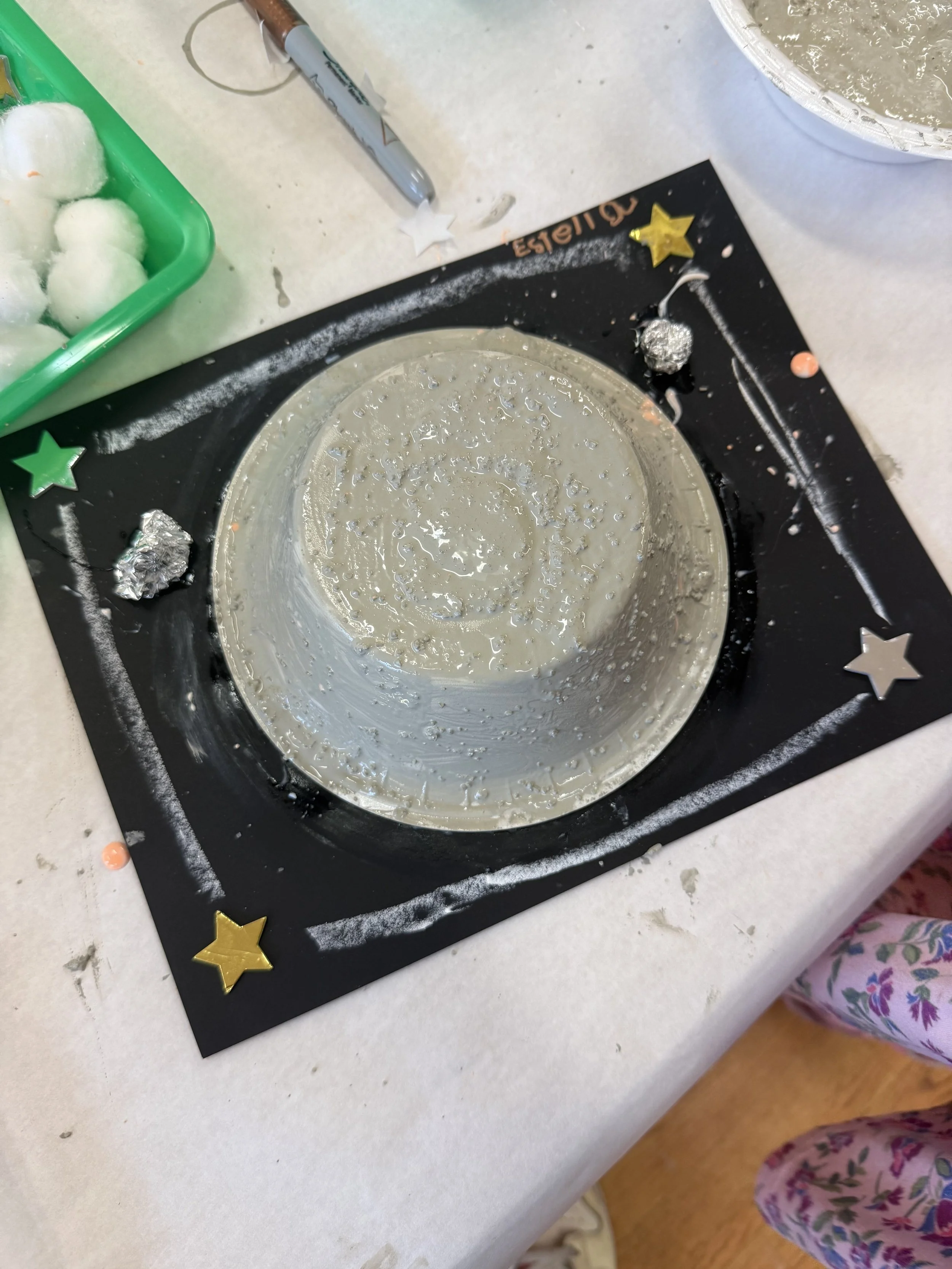 A round-shaped cake covered in white frosting, placed on a black board decorated with star-shaped stickers, and surrounded by craft supplies and decorations.