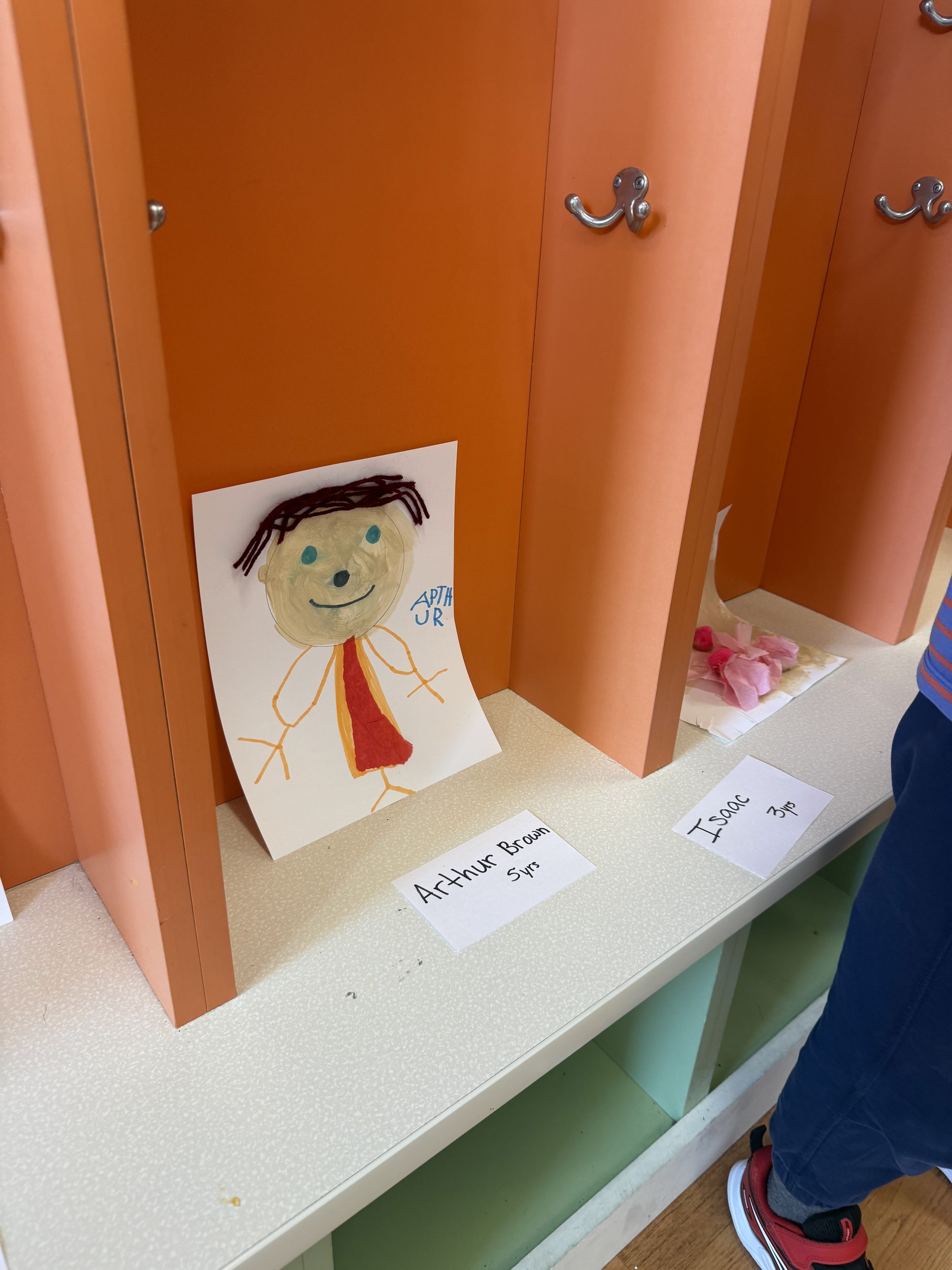 Child's drawing of a person with brown hair, tan face, and red shirt, labeled 'Arthur Brown 5 yrs' on a small white paper. The drawing is positioned outside a pink cubby in a classroom or daycare setting.